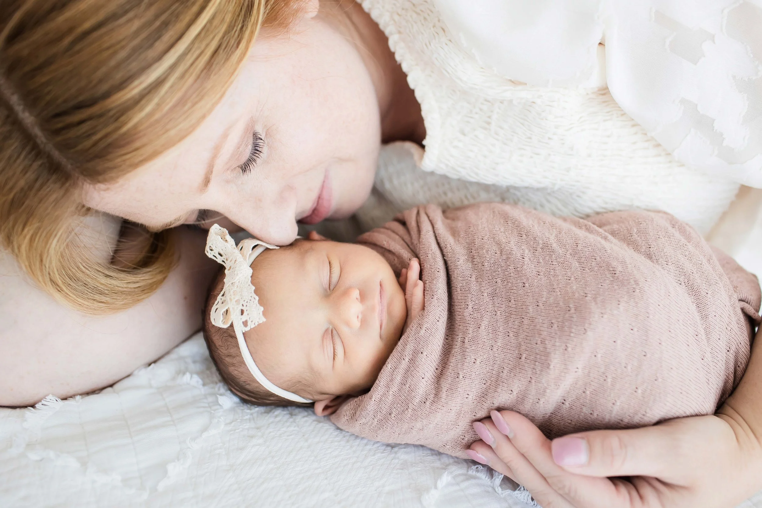 A woman with long blonde hair lying down and gently kissing a sleeping baby, both on a white textured blanket at East and Elm Portrait Studio in Fairport, NY, during a newborn session.