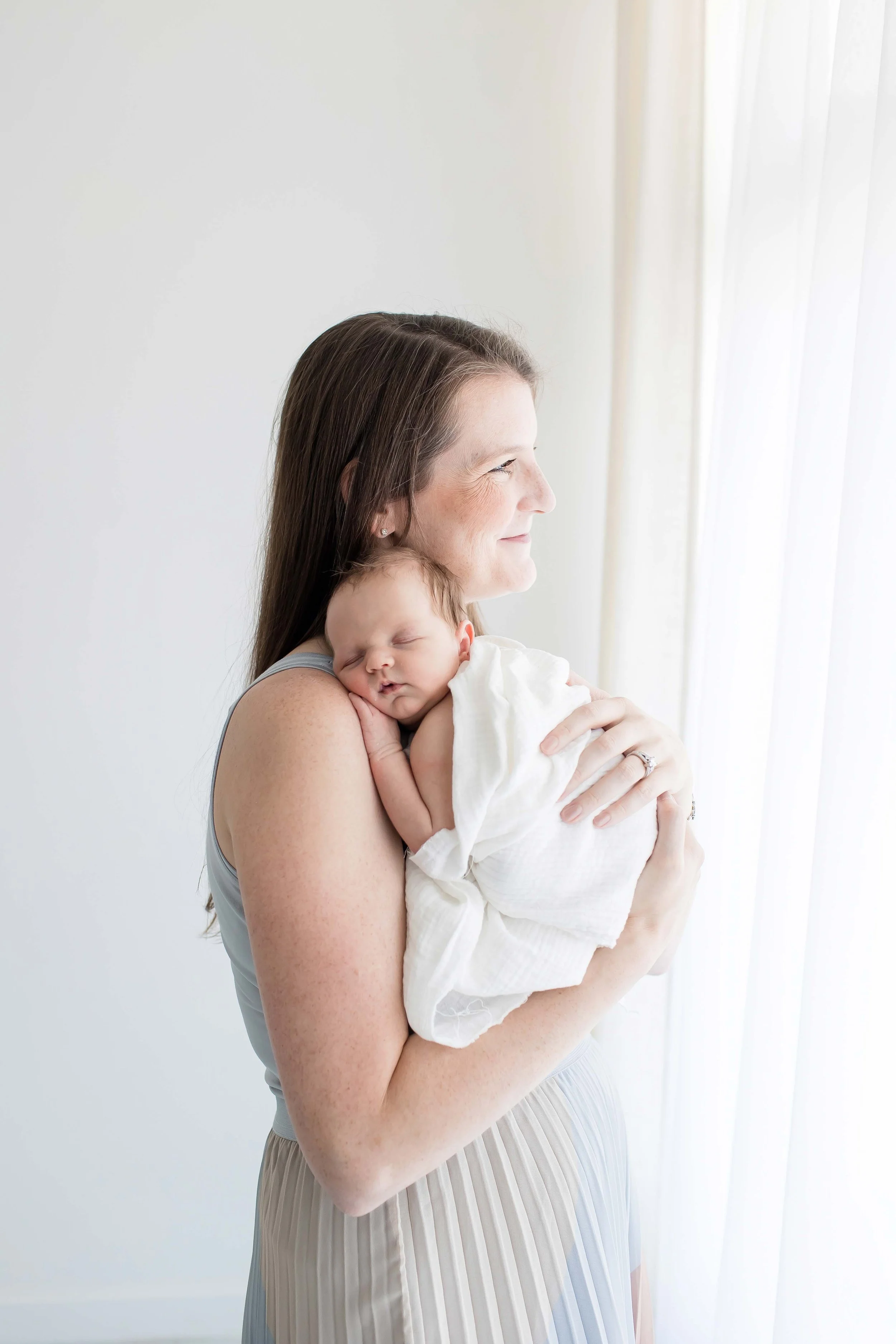 A smiling mom with long dark hair holding a sleeping baby wrapped in a white blanket near a window with sheer curtains at East and Elm Portrait Studio during a newborn session in Fairport, NY.