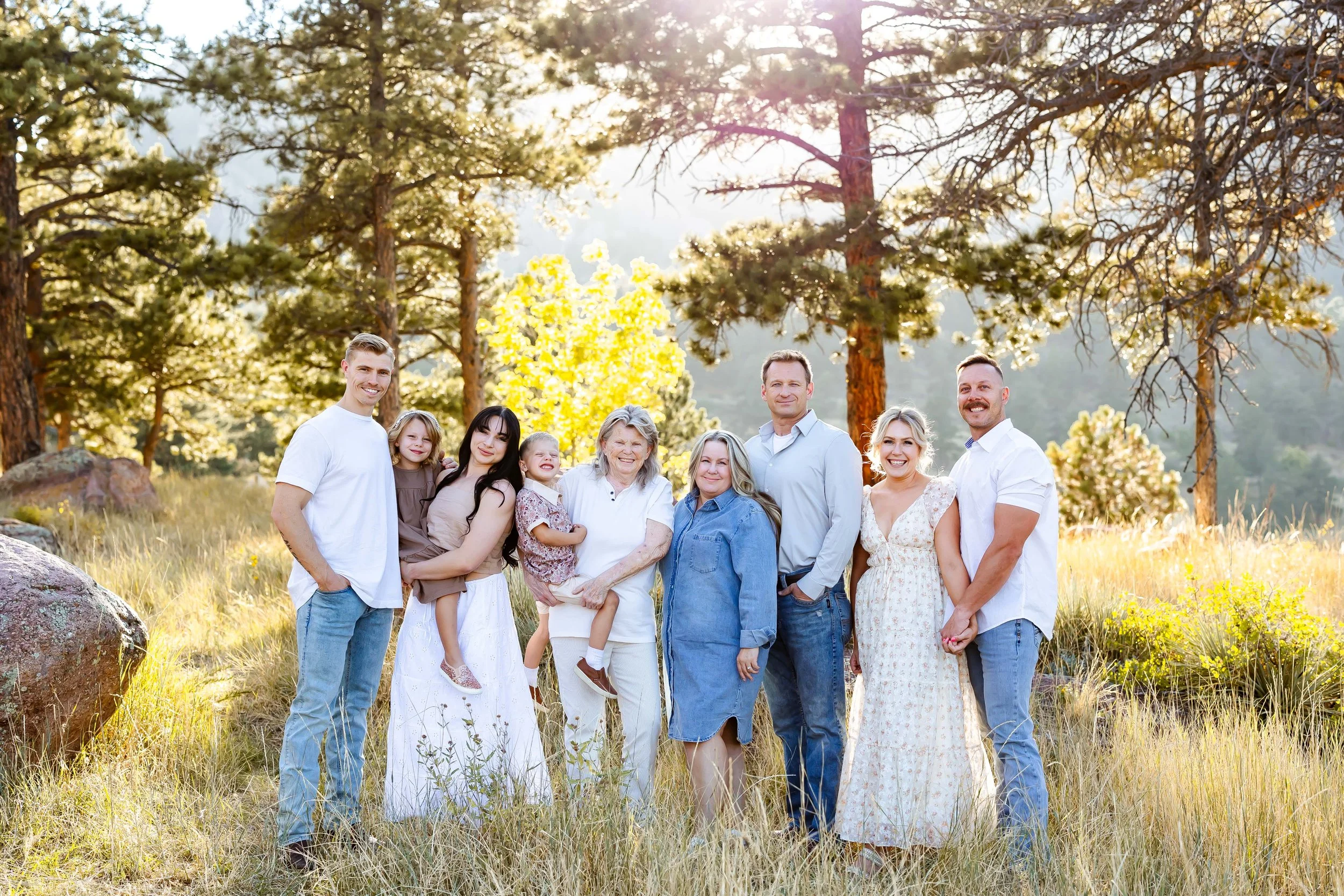 A large, multi-generational family posing outdoors in a forested area with tall trees and sunlight filtering through the leaves at a family session with East and Elm Portrait Studio in Rochester, NY.