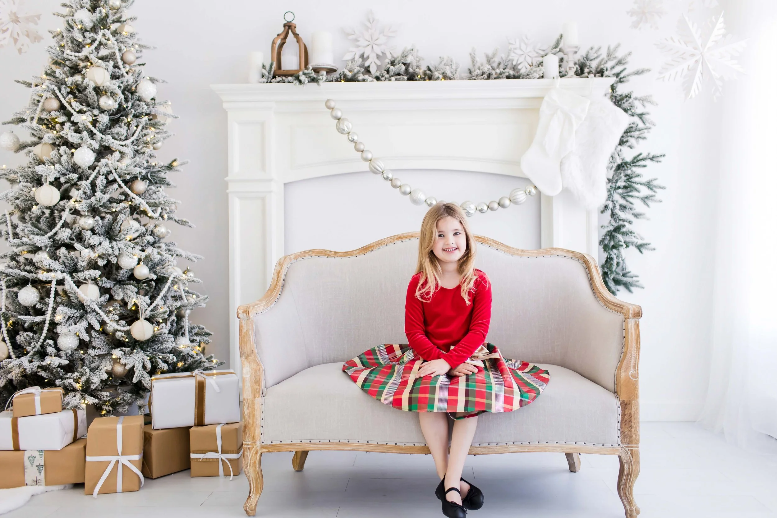 A young girl sitting in a Christmas-decorated studio, with a snow-flocked Christmas tree with ornaments at East and Elm Portrait Studio in Fairport, NY, during a Christmas mini session.