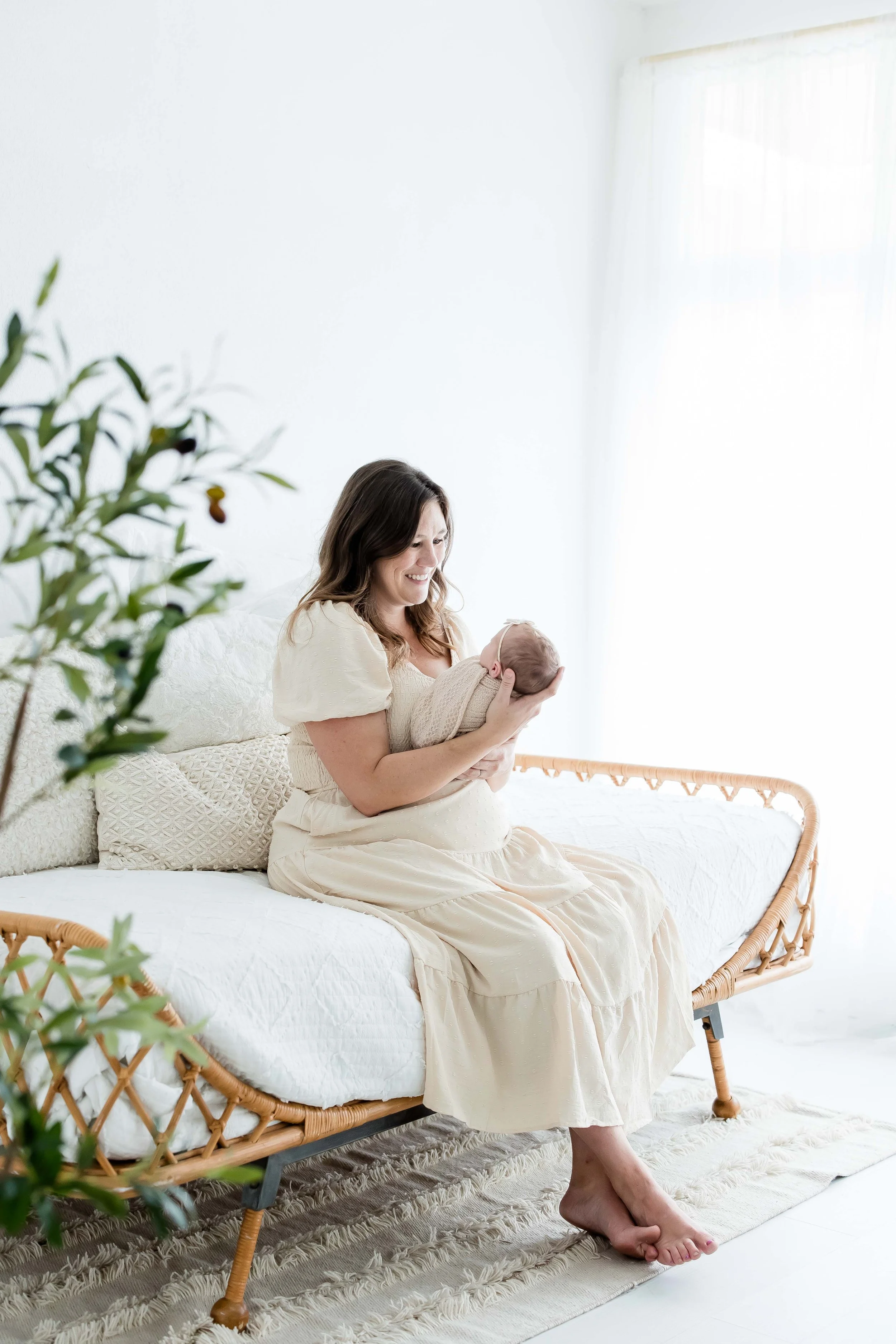 A woman sitting on a white bed holding a newborn baby in a beige blanket, at East and Elm Portrait studio in Fairport, NY.
