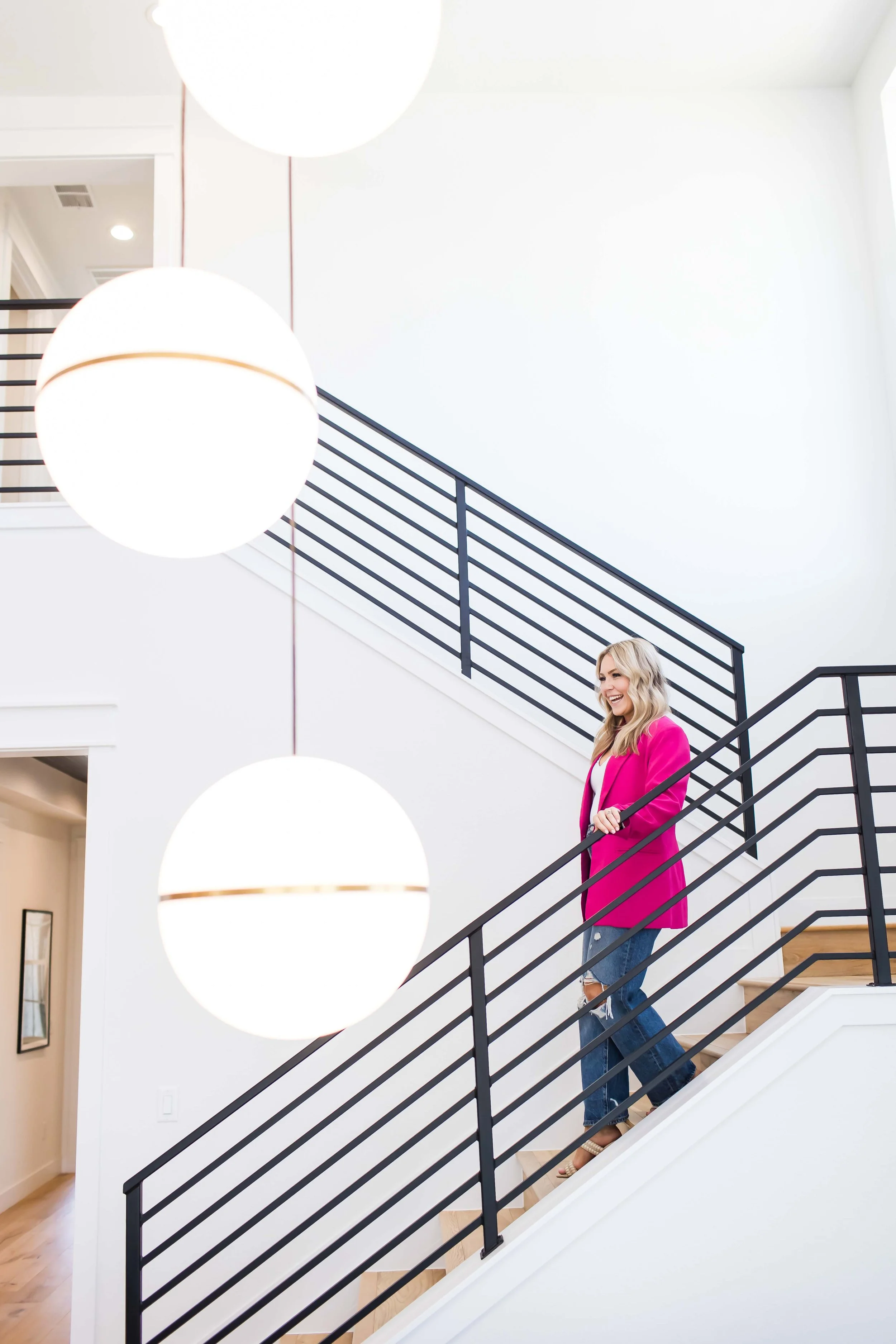 A woman with blonde hair wearing a bright pink blazer and ripped jeans walking down a staircase in a modern, minimalistic home interior during branding and headshot photos with East and Elm Portrait Studio in Rochester, NY.