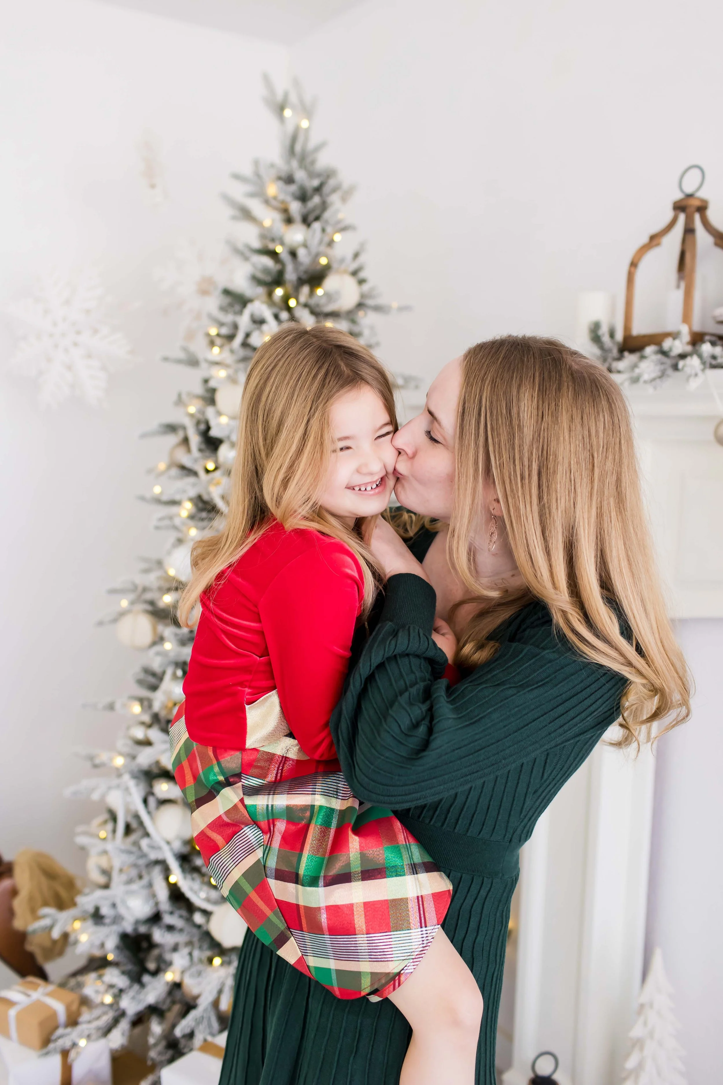 A mom and her daughter sharing a kiss in front of a decorated Christmas tree during Christmas mini sessions at East and Elm Portrait Studio in Fairport, NY