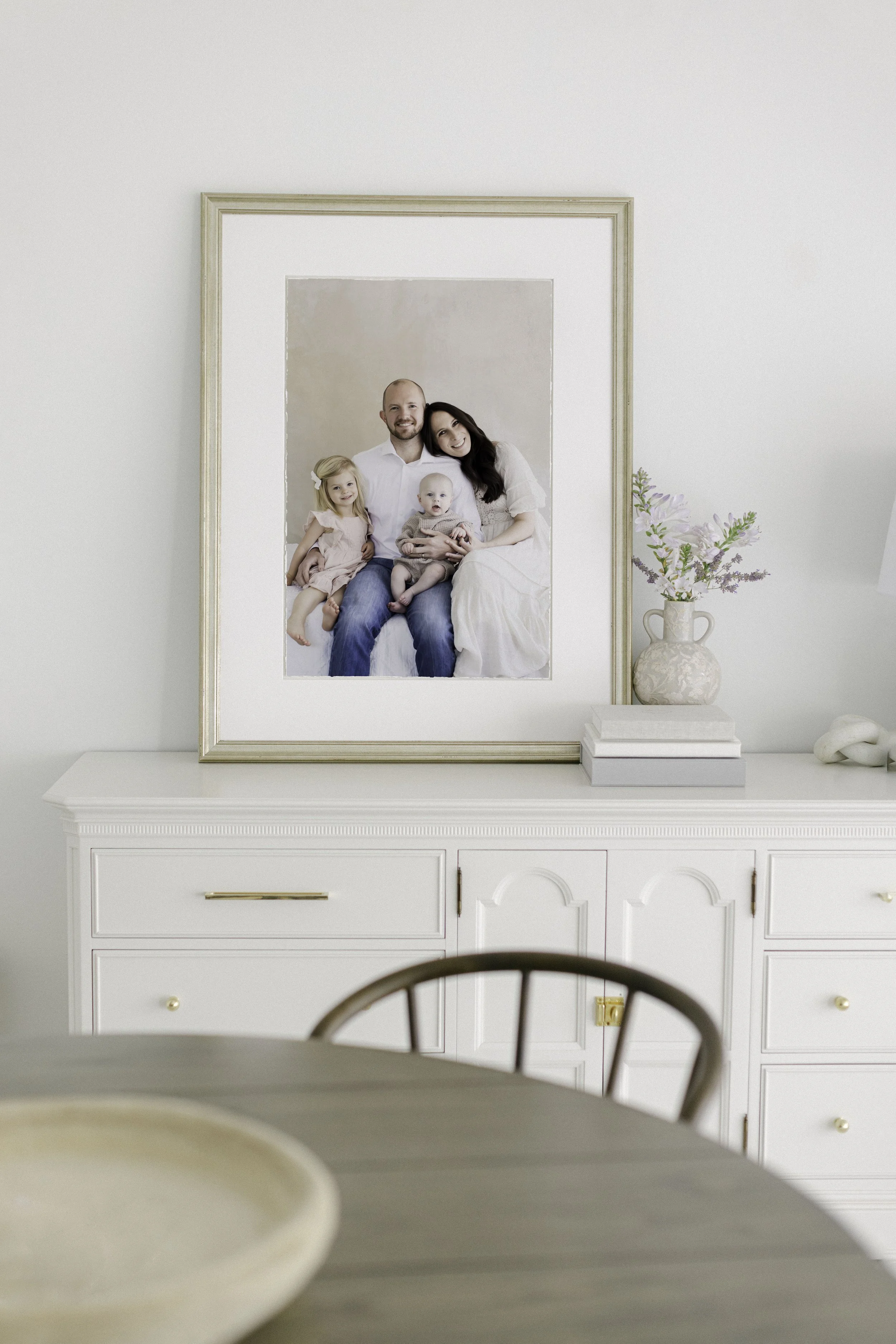 A framed family portrait on a white sideboard, with a vase of flowers beside it, in a minimalist background.