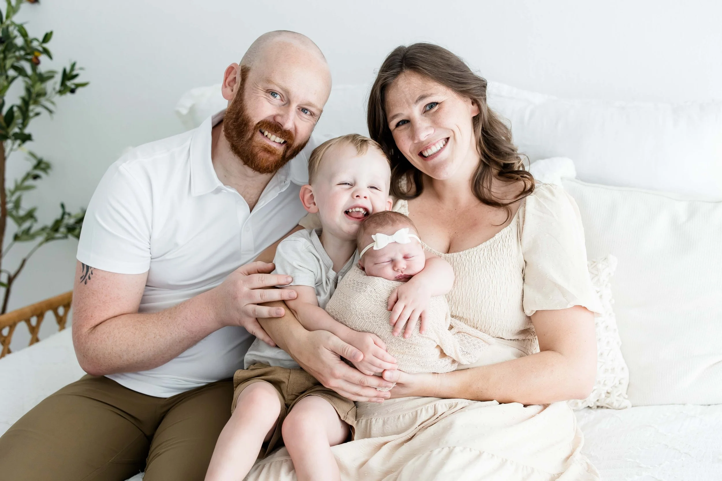 A happy family of five sitting on a bed with a white background at East and Elm Portrait Studio in Fairport, NY, during a newborn session.