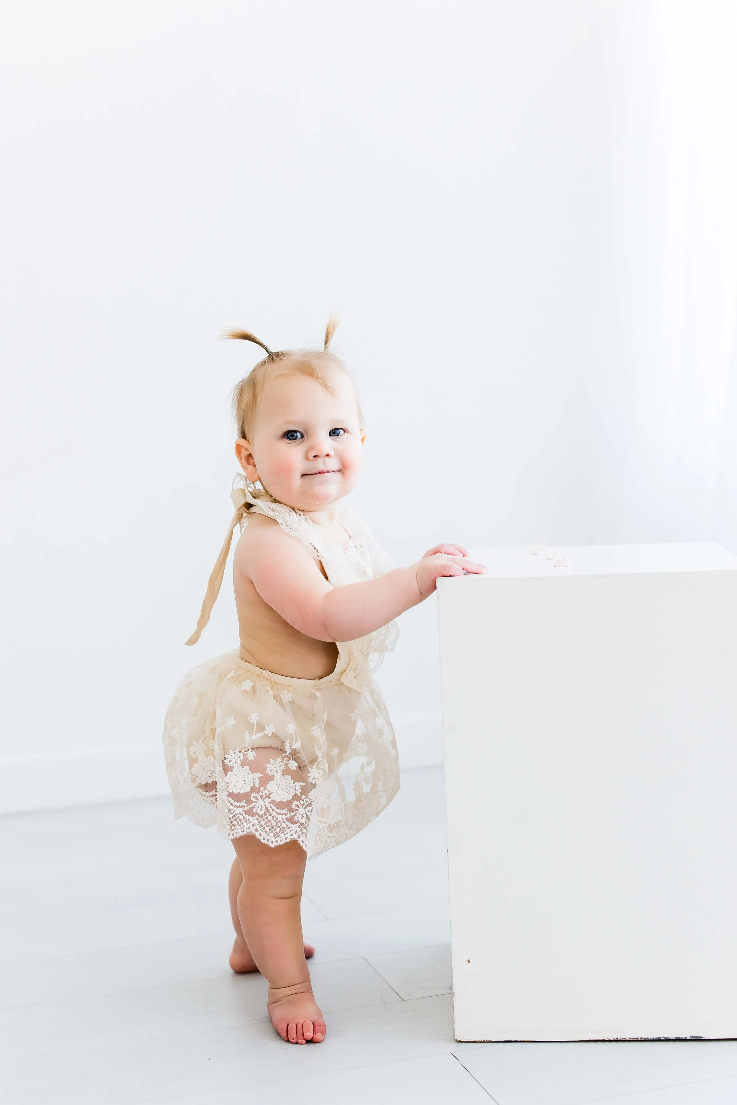 A young girl with blonde hair standing next to a white rectangular block at East and Elm Portrait Studio during a studio child's photo session in Fairport, NY.