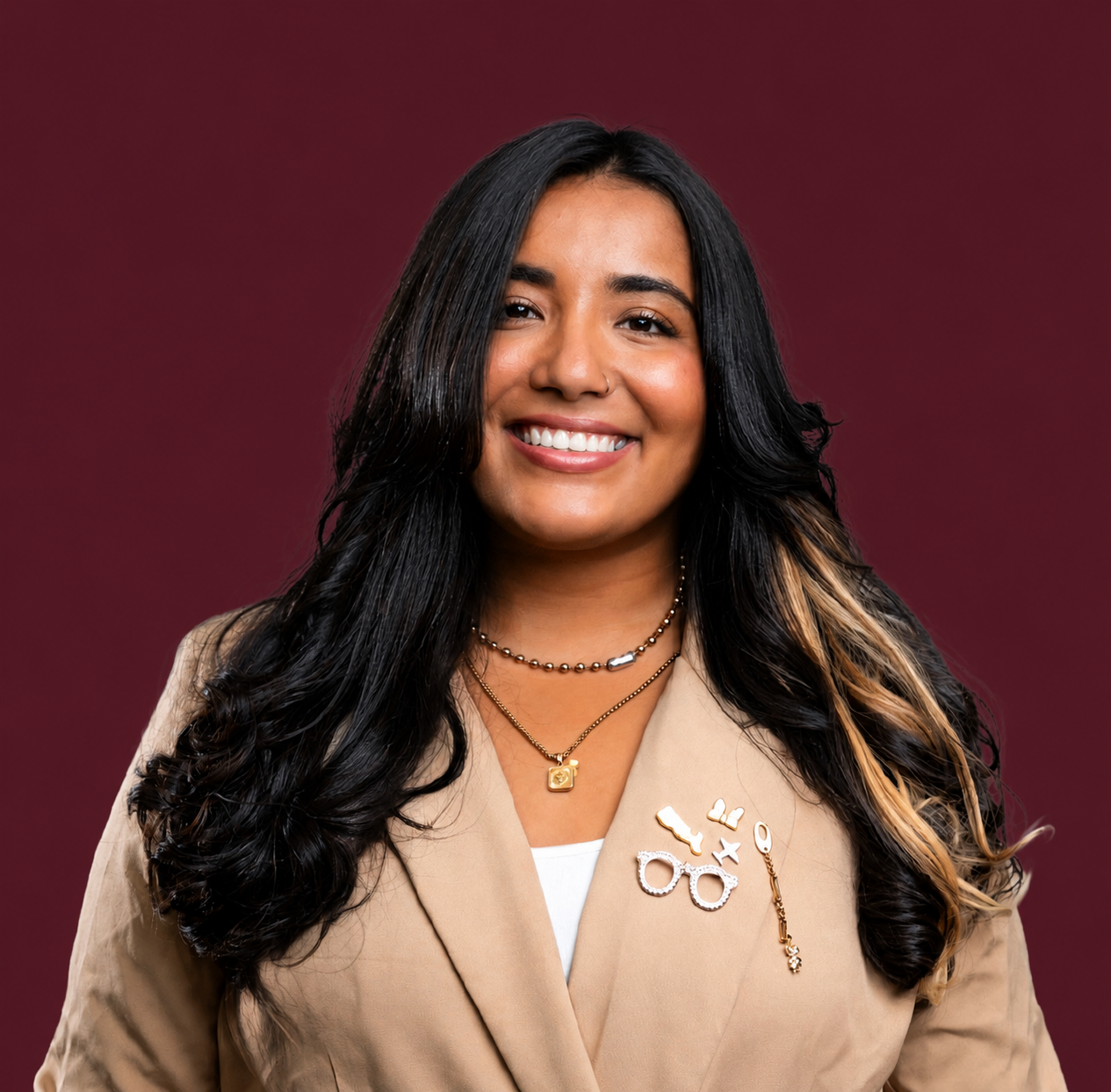 A woman with long, dark, wavy hair smiling, wearing a beige blazer adorned with pins and necklaces, against a maroon background.