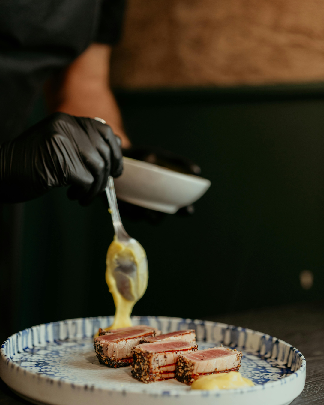 A chef wearing black gloves is serving slices of seared tuna topped with a soft-cooked egg yolk on a patterned plate.