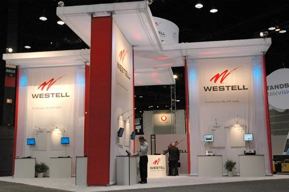 Trade show booth for WESTELL with white and red design, multiple screens and employees attending to visitors.