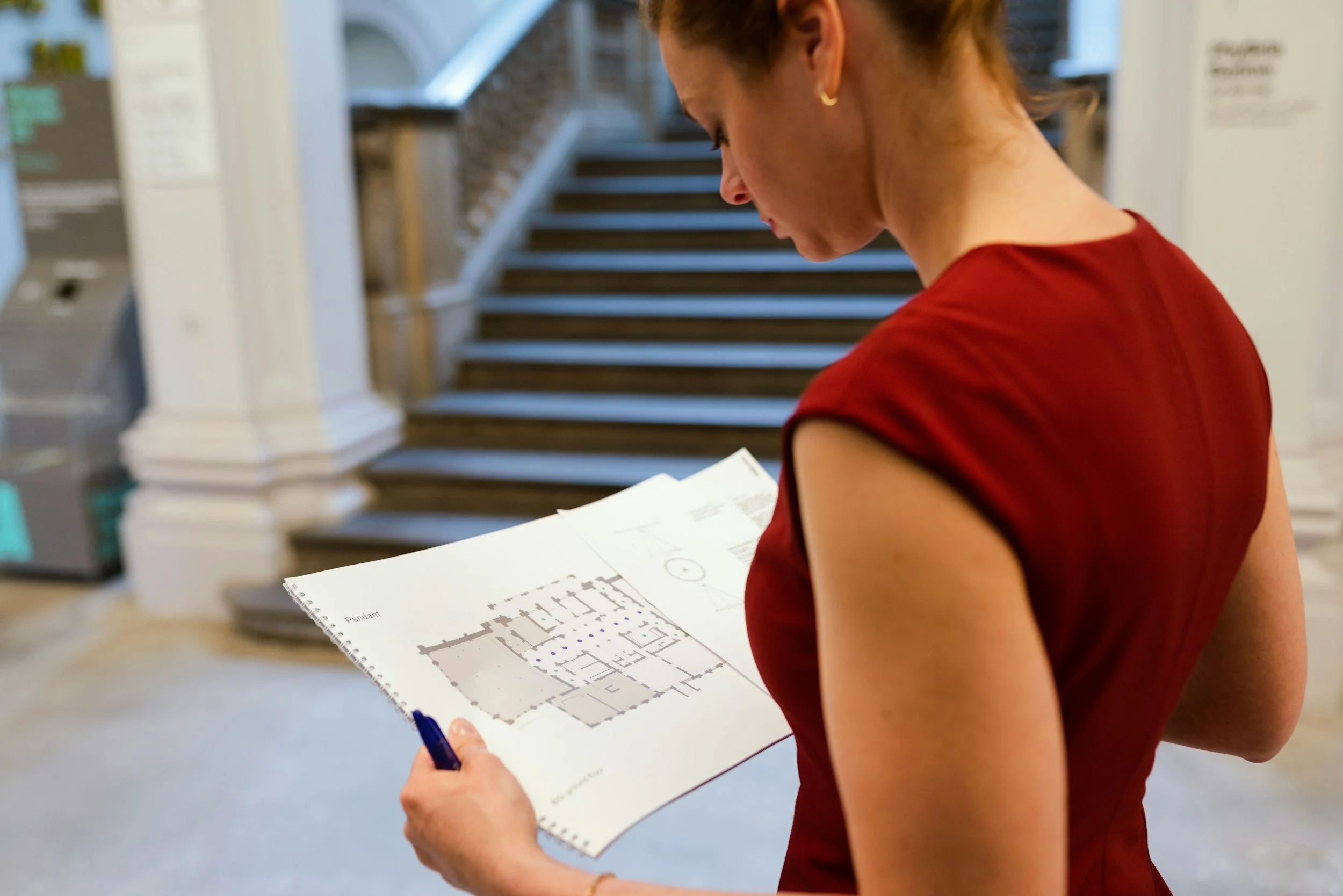 A woman in a red sleeveless top is reading an architectural floor plan and holding a pen, in a building with a staircase in the background.