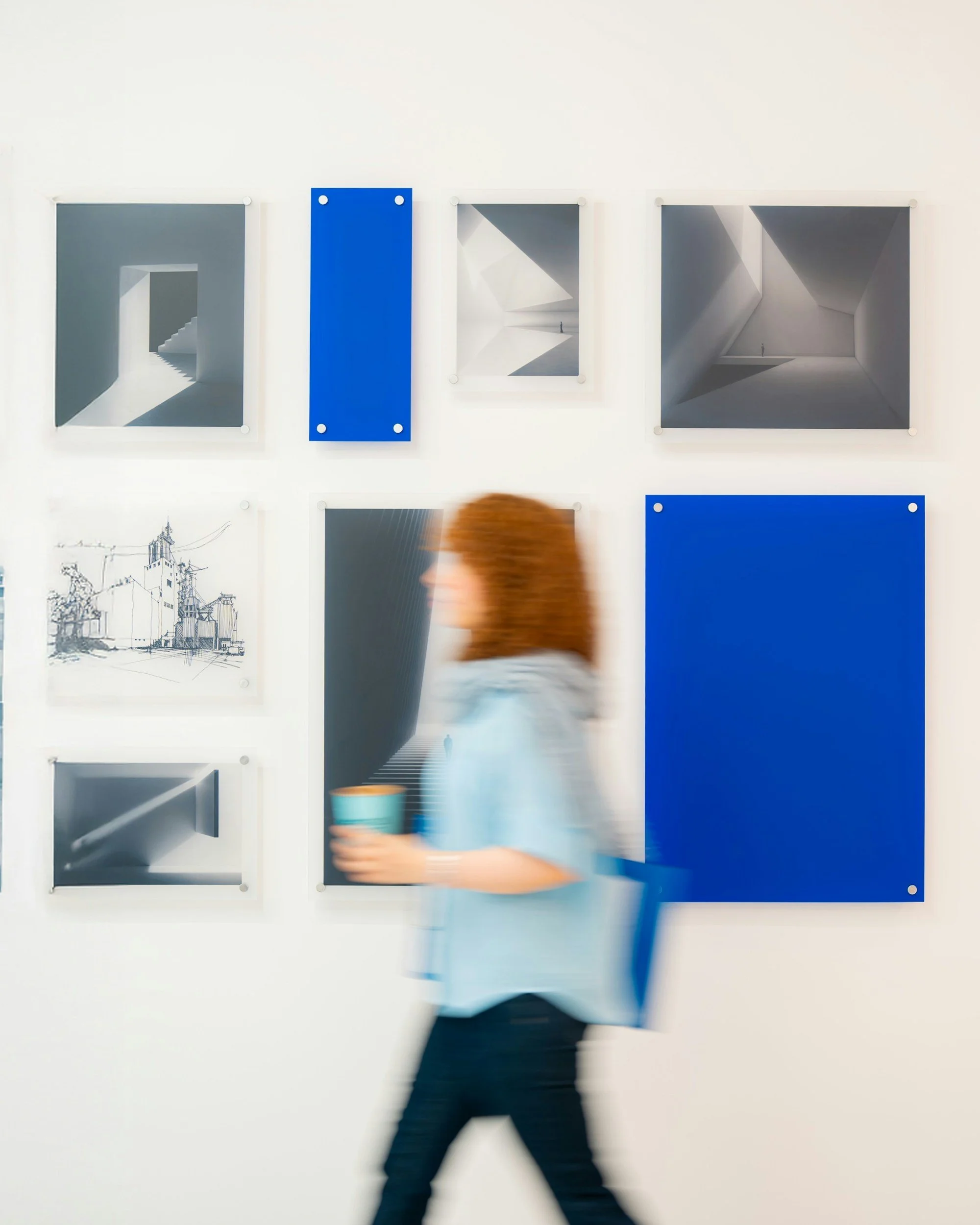 Blurred person walking past a white wall with framed black-and-white artwork, some blue artwork, and a drawing of buildings.