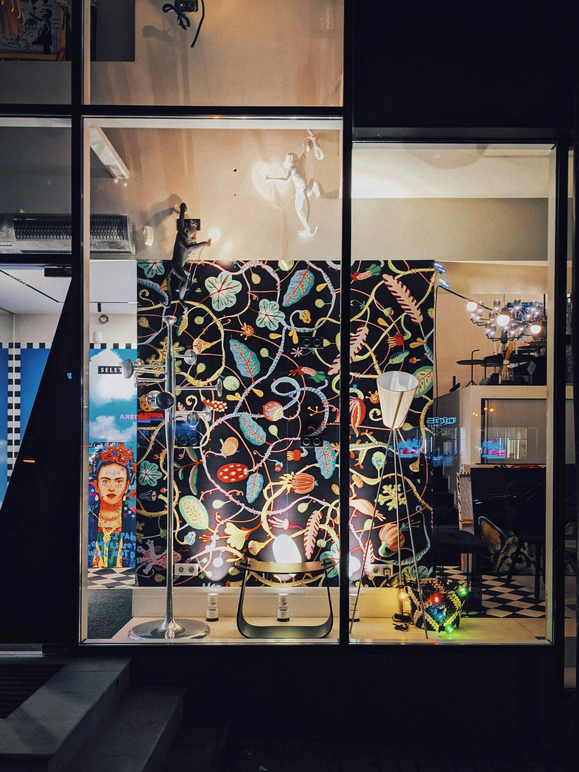 A reflection of a storefront window showing a colorful, intricate floral and leaf mural, with various objects inside including a vintage lamp, a person with a camera taking a photo, and a framed portrait of Frida Kahlo.