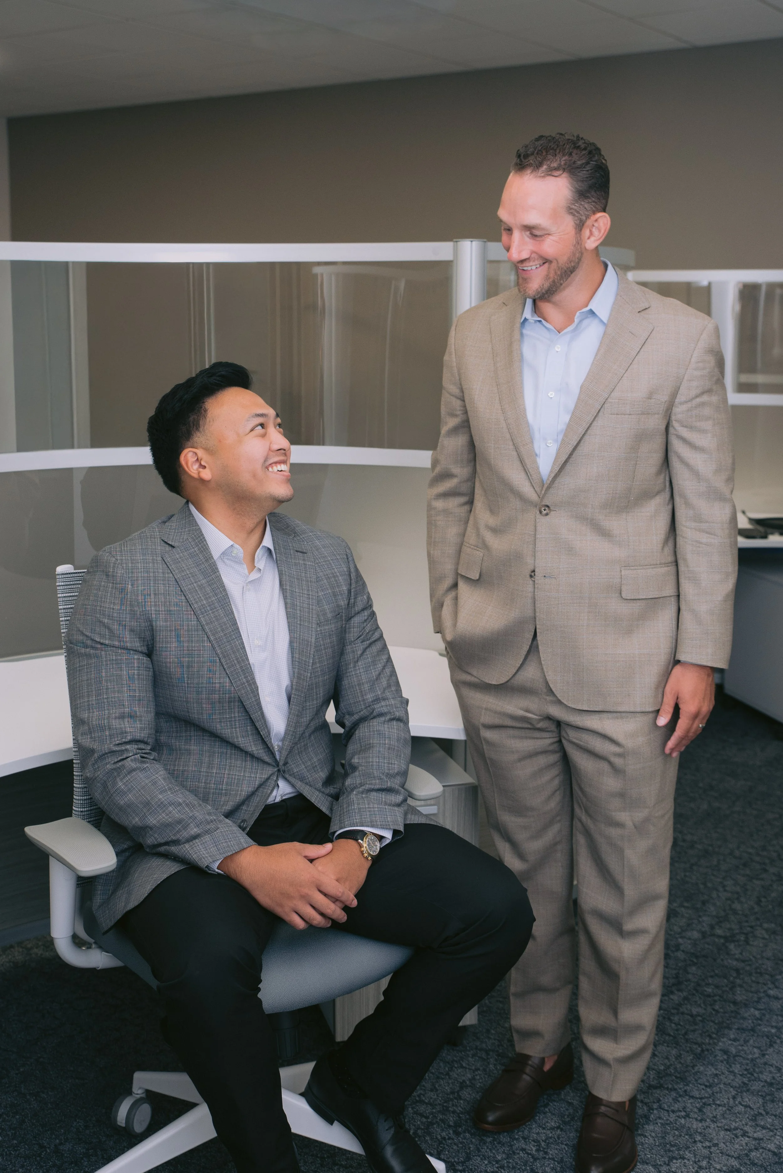 Two men in business attire engaging in conversation in an office environment. One man is sitting in a grey office chair, smiling, while the other man is standing and smiling at him.