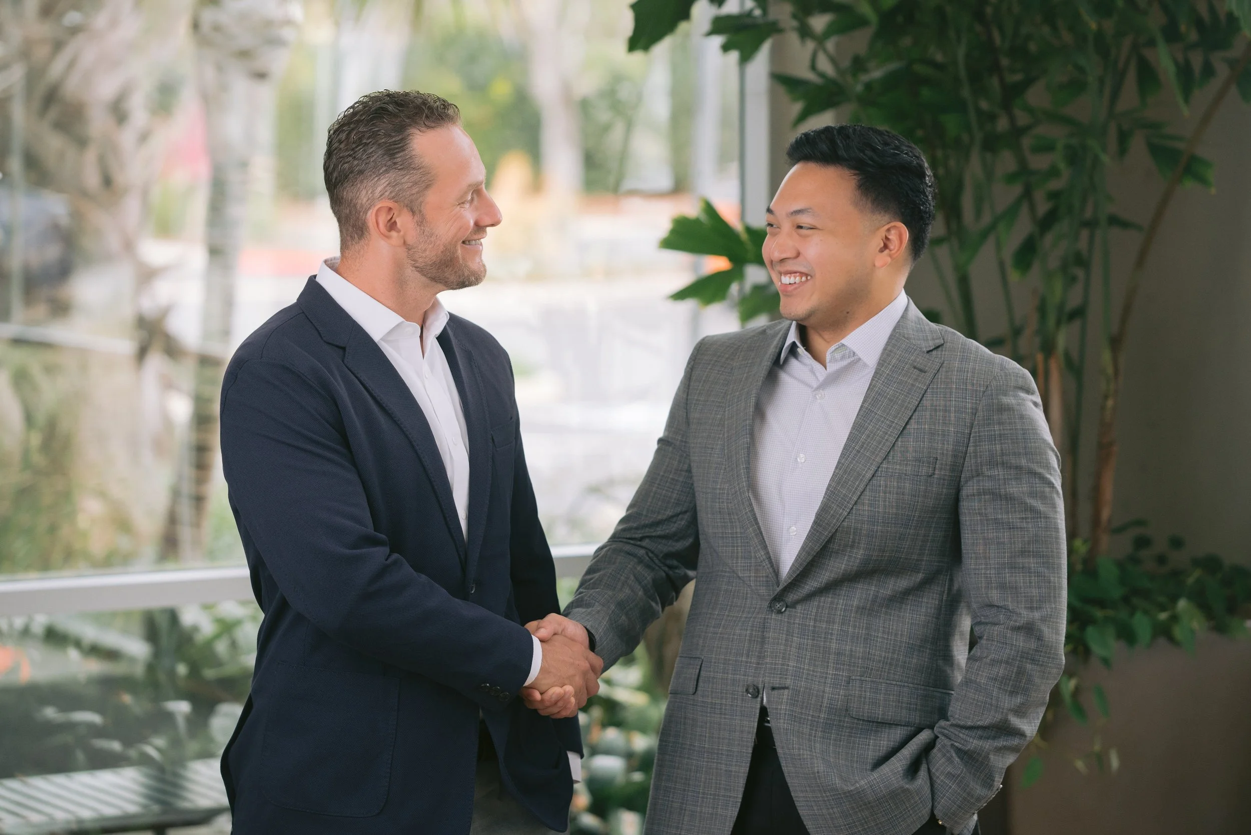 Two men in business suits shake hands and smile at each other indoors with large windows and green plants in the background.