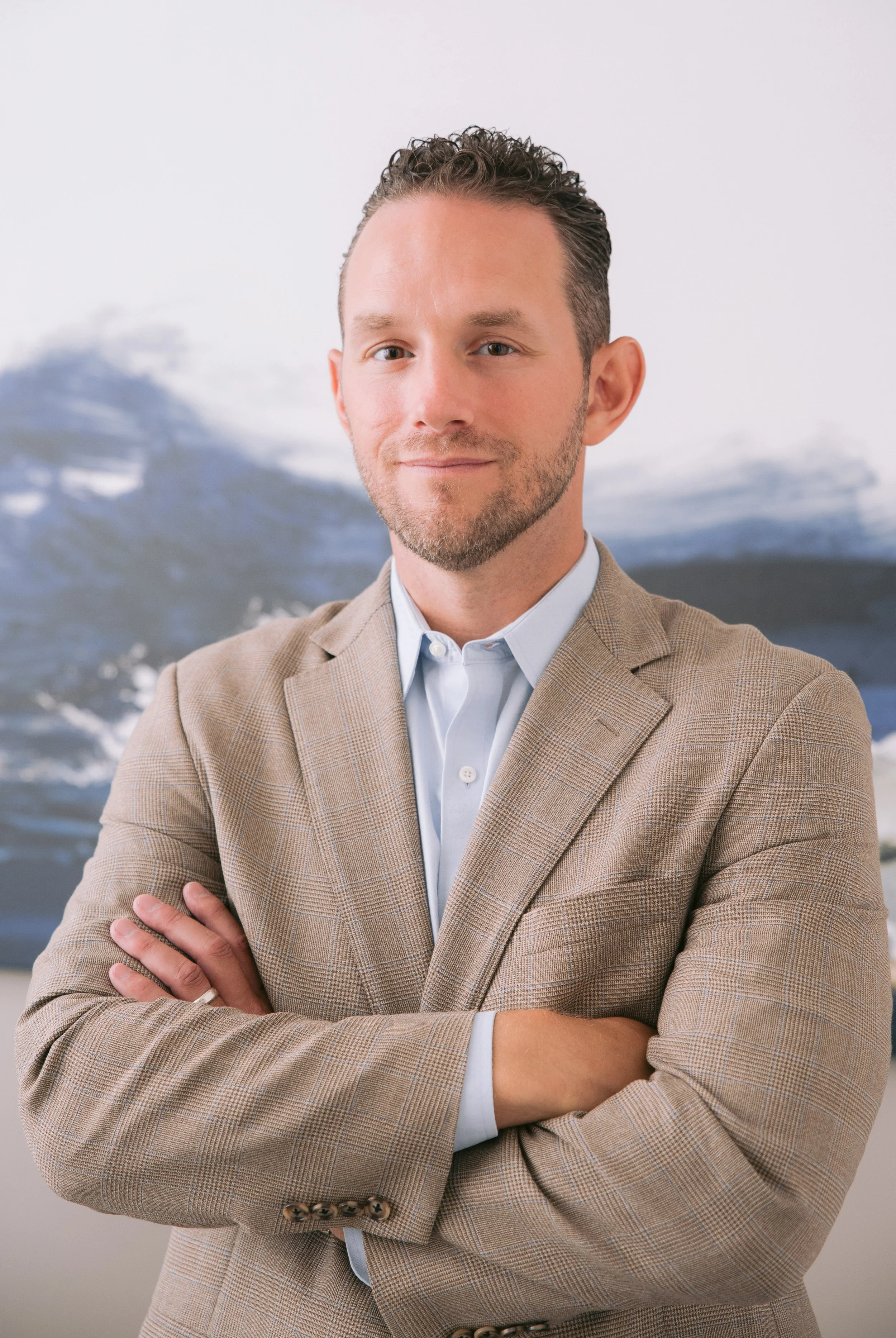 A man with short, curly hair, light skin, and a beard, wearing a beige checked blazer and a light blue shirt, stands with his arms crossed in front of a backdrop of snow-capped mountains.