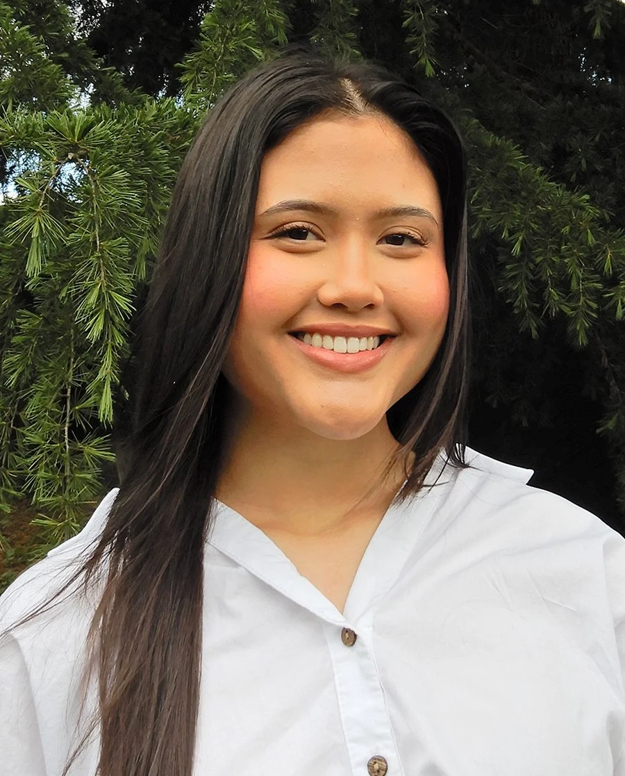 A young woman with long dark hair smiling outdoors in front of greenery.