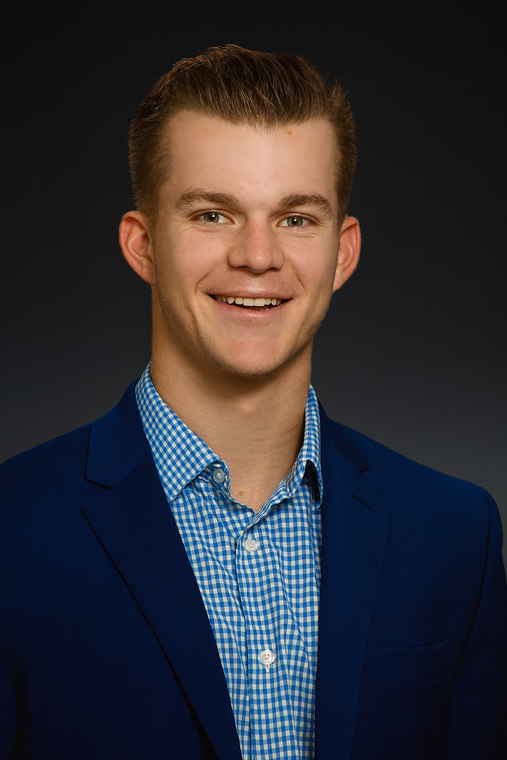 Headshot of a young man with blonde hair, smiling, wearing a blue blazer over a blue and white checkered shirt, against a dark background.