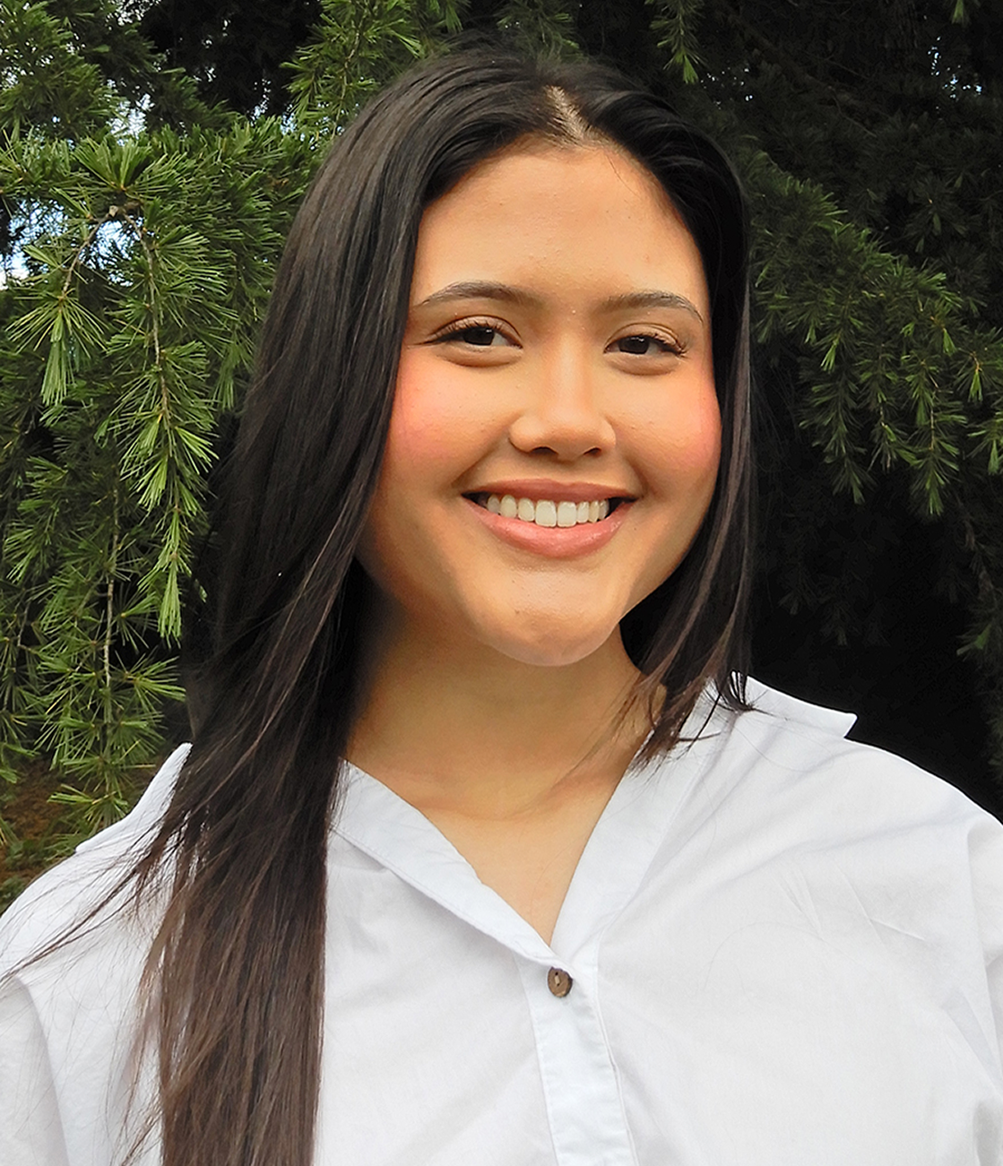 A smiling woman with long dark hair posing outdoors in front of green foliage, wearing a white shirt.