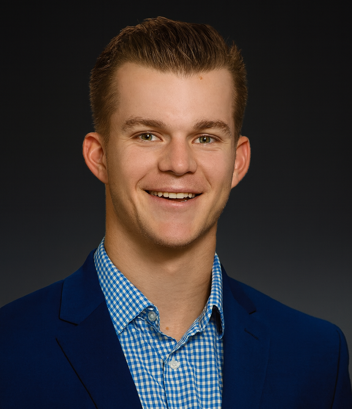 Young man with short brown hair, wearing a blue suit jacket and a blue checkered shirt, smiling against a dark background.