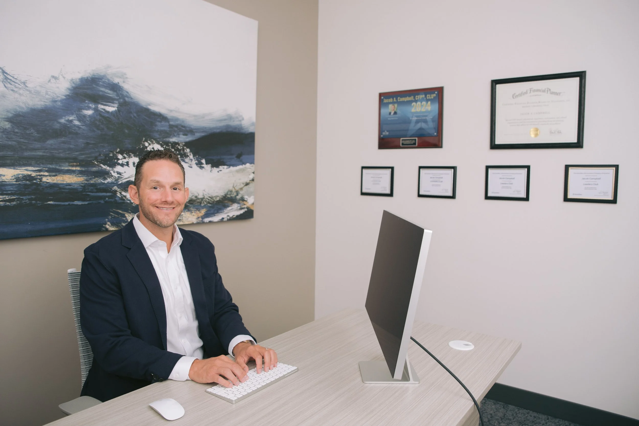 A man in a business suit sitting at a desk with an all-in-one computer, typing on a wireless keyboard, in an office with certificates and awards on the wall, and a large ocean-themed painting.