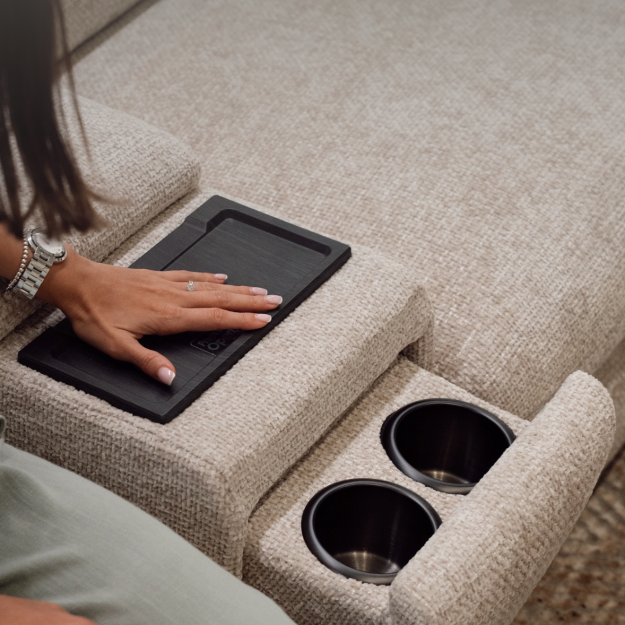 A person wearing a watch and jewelry placing a rectangular black object on a beige carpeted surface near two black cylindrical containers.