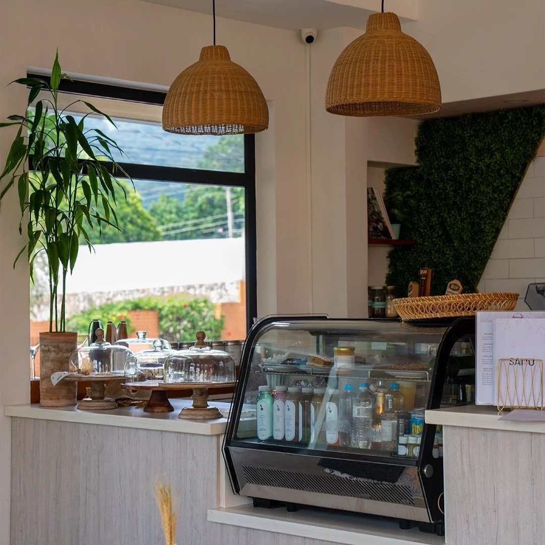 Interior of a cafe with a counter displaying pastries under glass domes, a refrigerated display case with bottled drinks, a large window showing outdoor greenery, and decorative hanging lamps.
