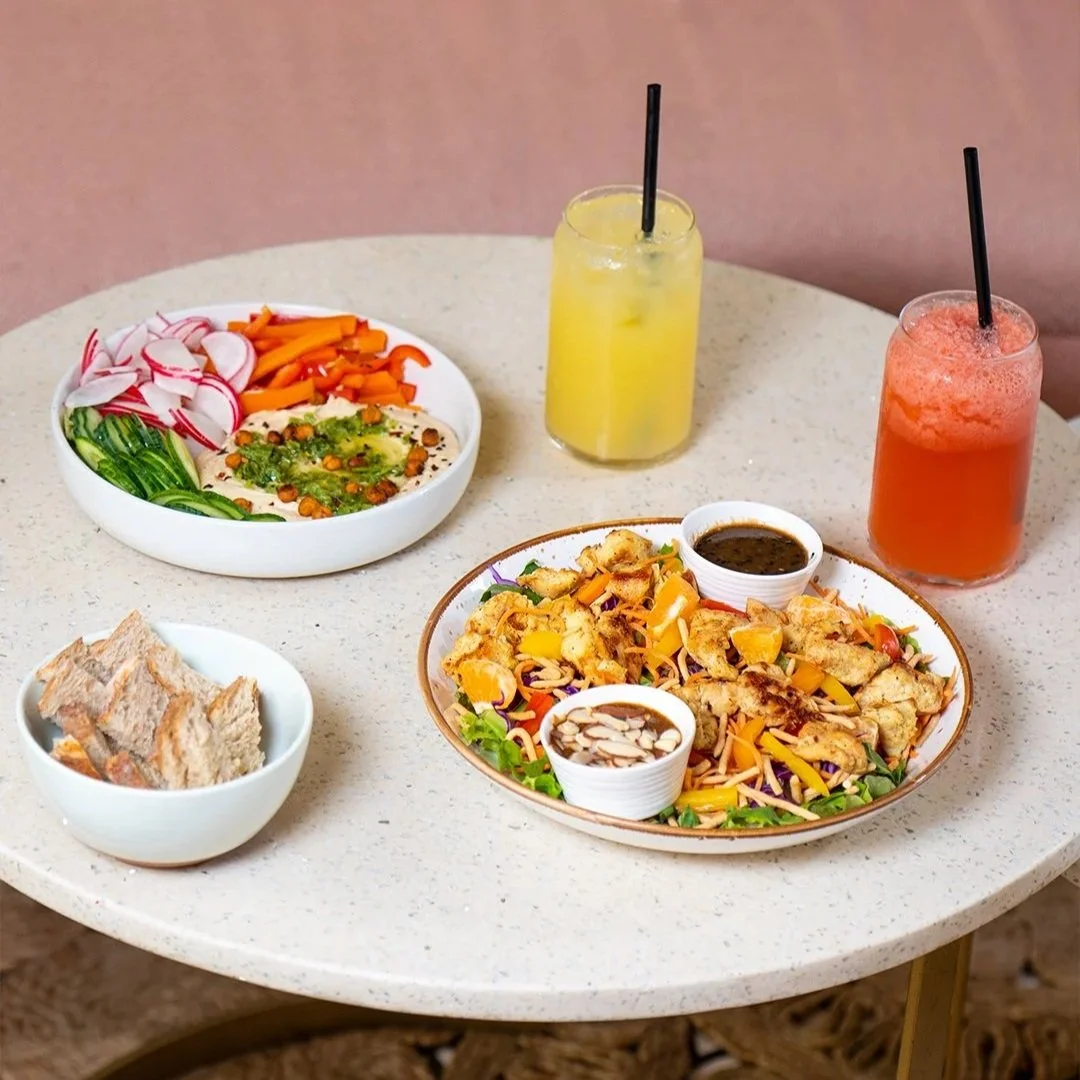 A round table with a salad, a chicken and vegetable dish, two drinks, and a bowl of bread. The salad includes radishes, carrots, and cucumbers. The chicken dish has shredded chicken, shredded cheese, and two small bowls of sauce and sliced almonds.