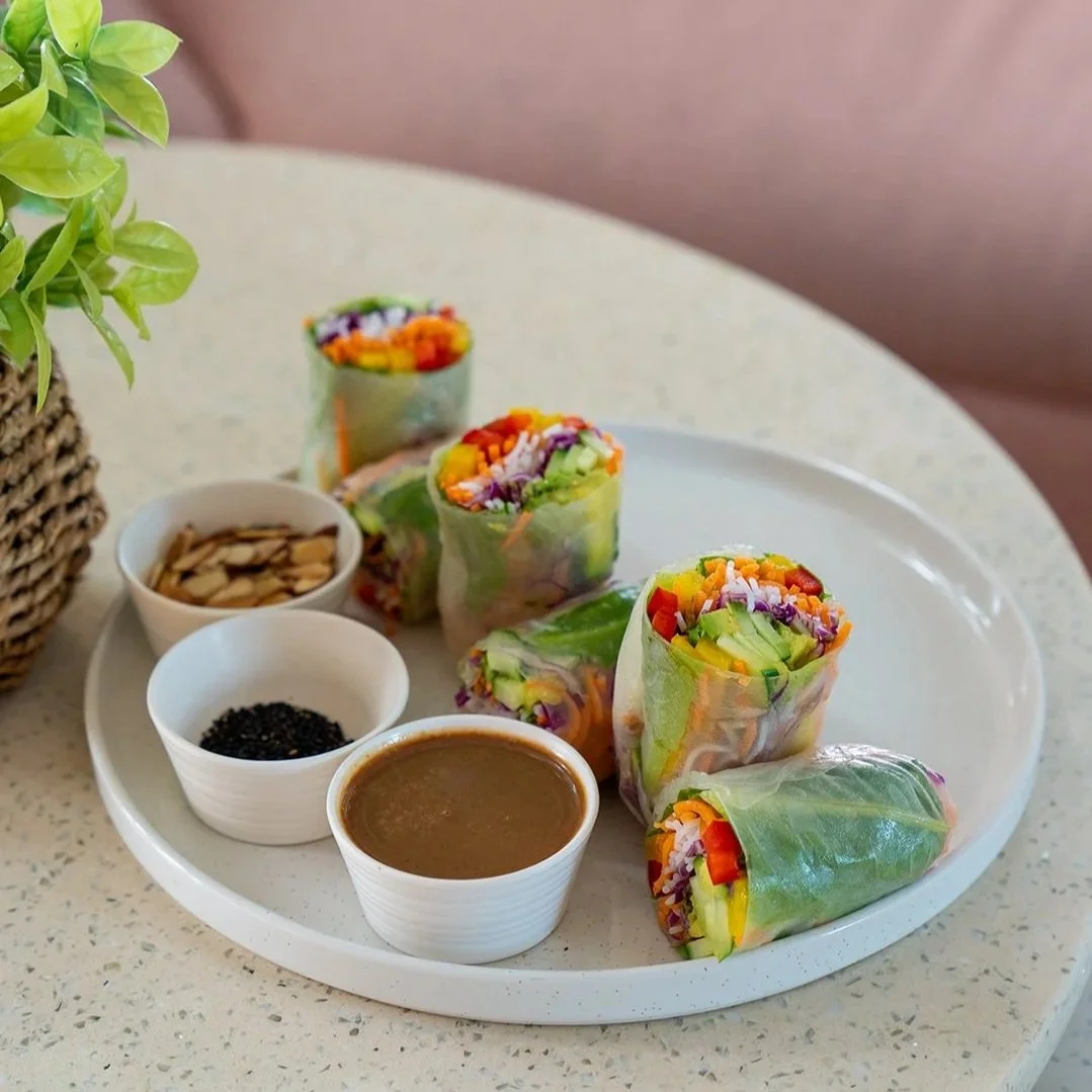 Colorful vegetable spring rolls on a white plate with dipping sauces, almond slices, and black sesame seeds, next to a potted green plant on a round table.