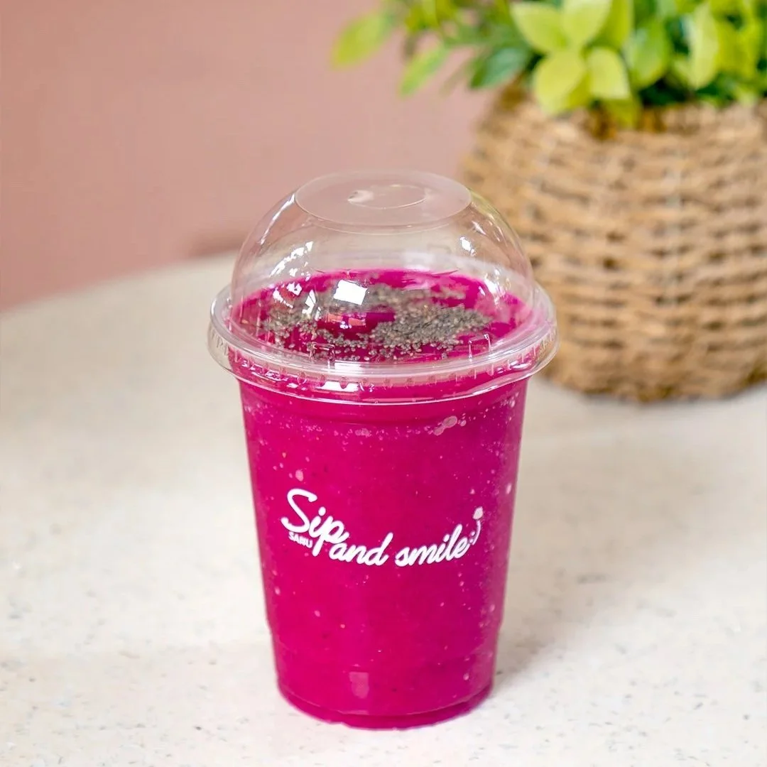 A bright pink smoothie in a clear plastic cup with a dome lid, sitting on a white surface with a blurred plant in a basket in the background.