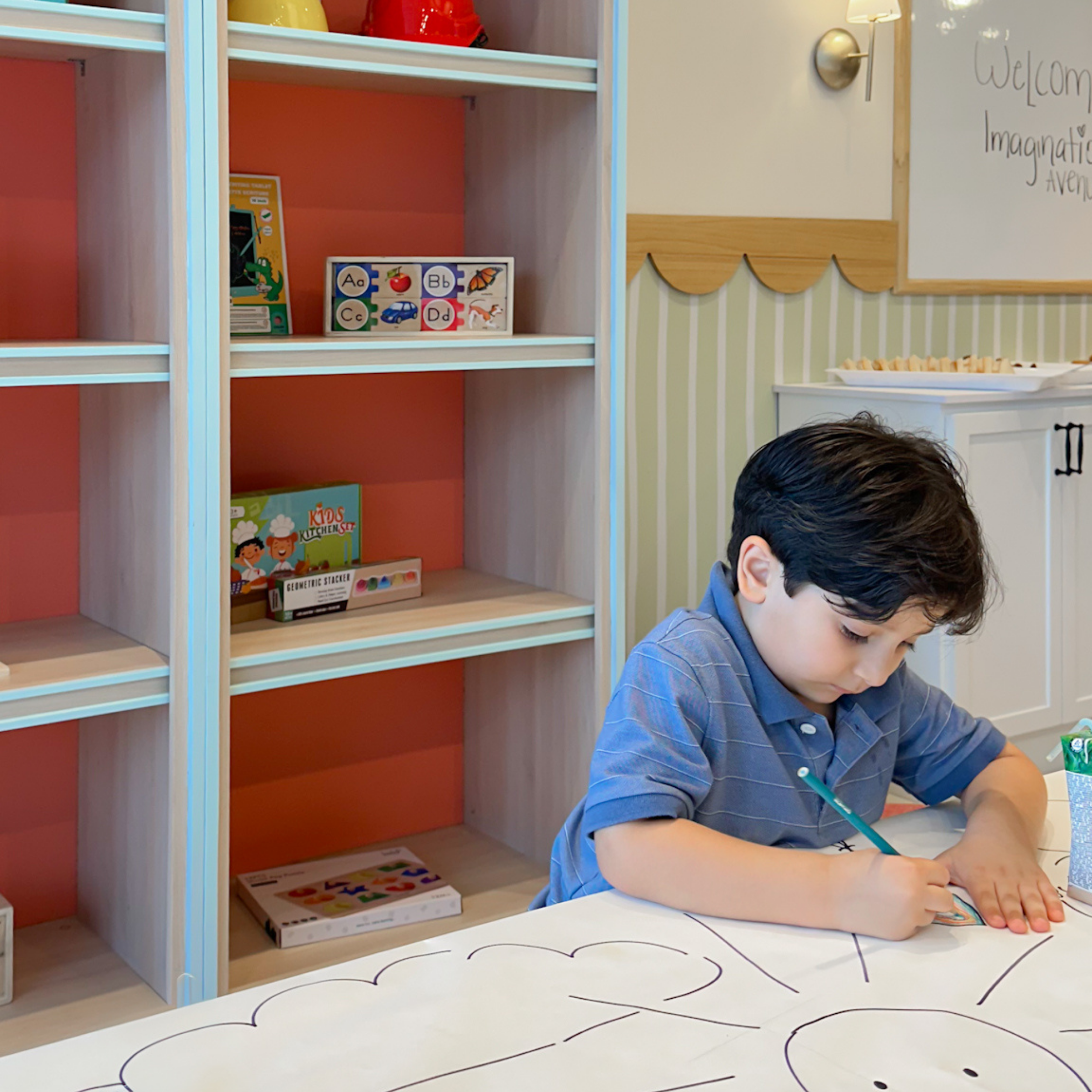 A young boy with dark hair wearing a blue shirt is sitting at a white table with black markings, working on a craft or activity. Behind him is a wooden shelf with books and educational toys, and to his right, a decorated wall with a whiteboard and a 