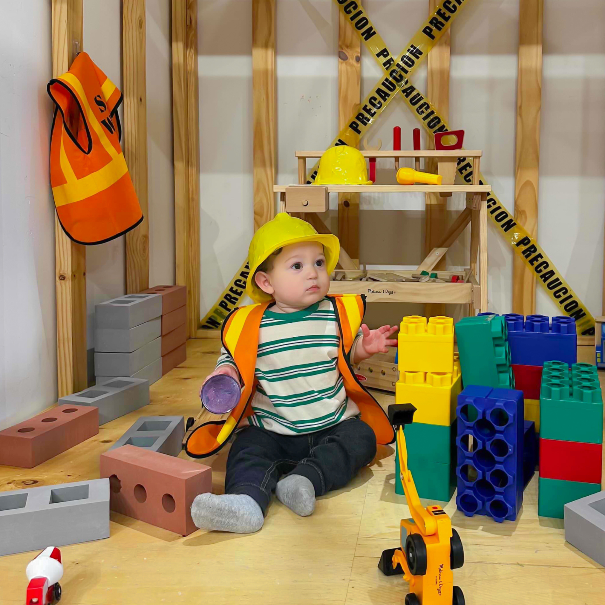 A young child wearing a yellow construction hard hat and a construction vest, sitting on a wooden floor surrounded by colorful building blocks, with a wooden construction backdrop and caution tape in the background.