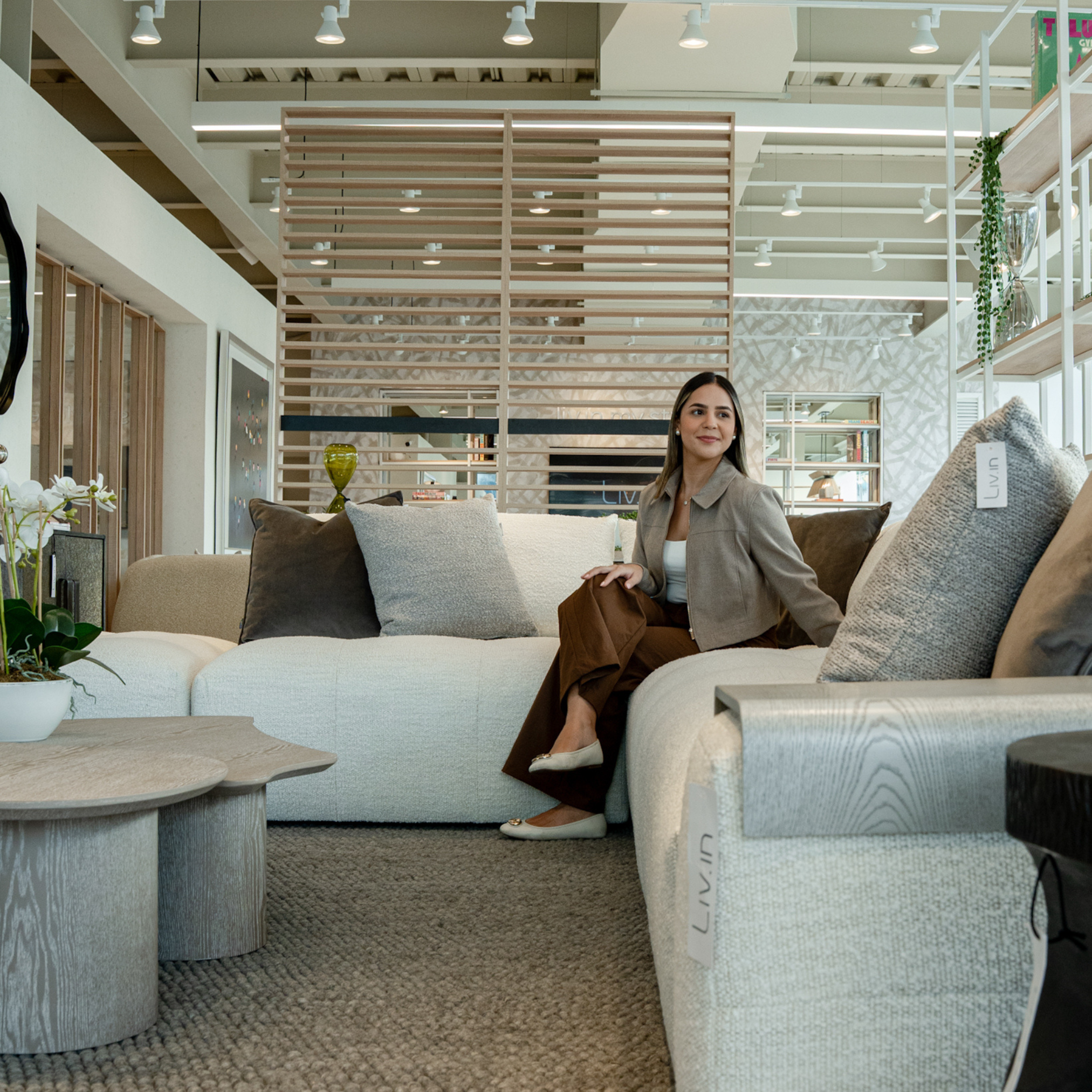 A woman sitting comfortably on a sofa in a modern, well-lit furniture store, with decorative pillows and a wooden coffee table in front of her.