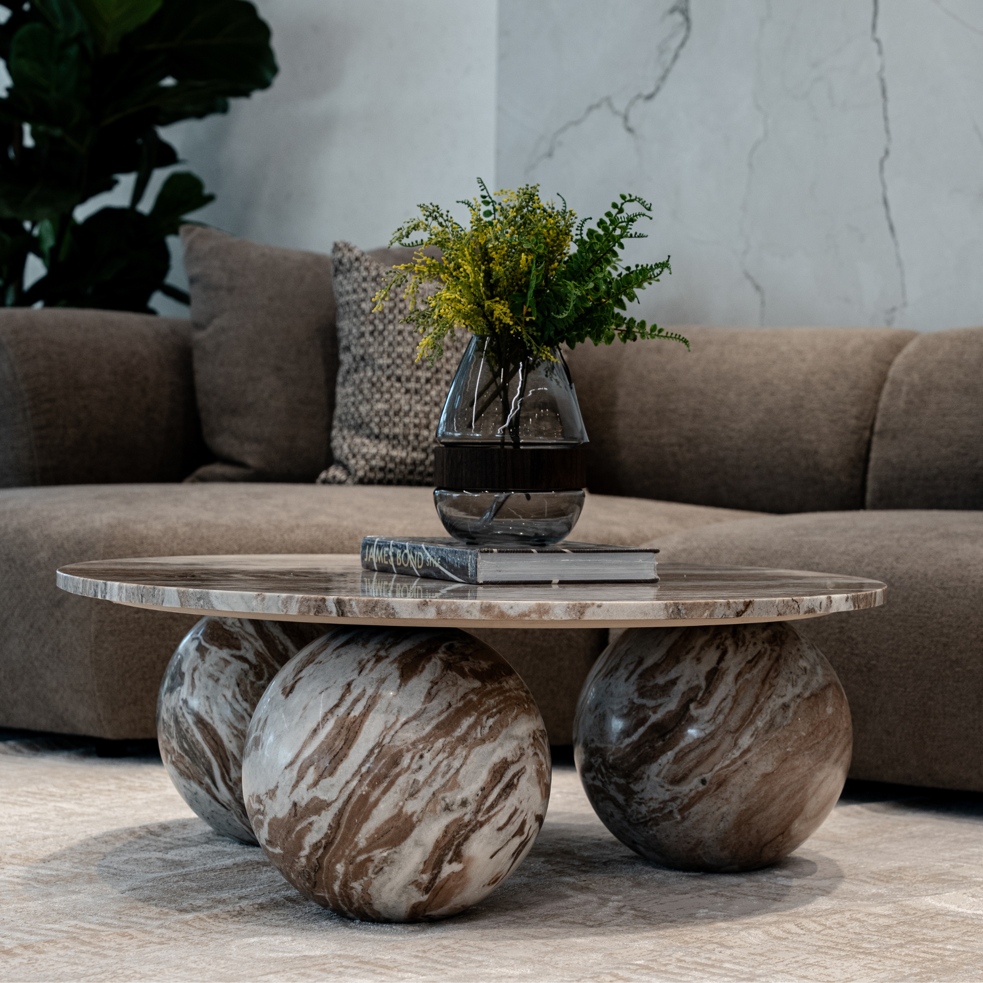 A glass vase with a bouquet of yellow and green foliage on a table in front of a beige sofa with patterned pillows, in a living room with a textured wall.