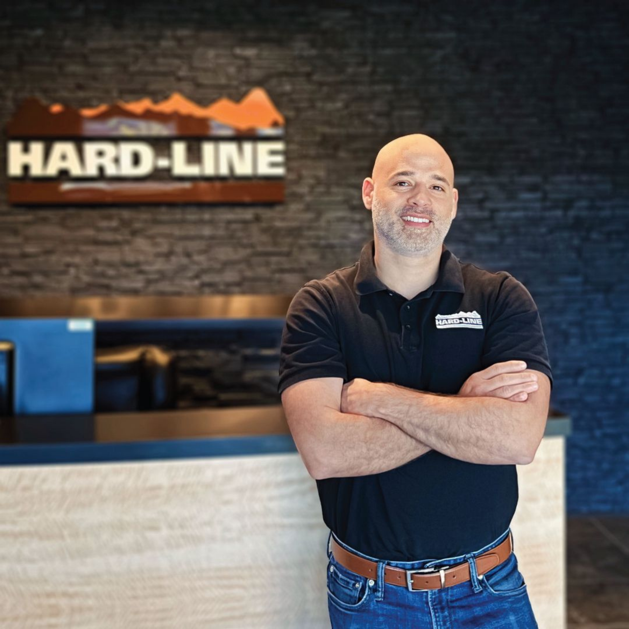 A smiling man with a shaved head and light stubble, wearing a black polo shirt with a logo that says 'HARD-LINE' on it, smiling with his arms crossed in front of a textured dark gray brick wall with a 'HARD-LINE' logo sign in the background.