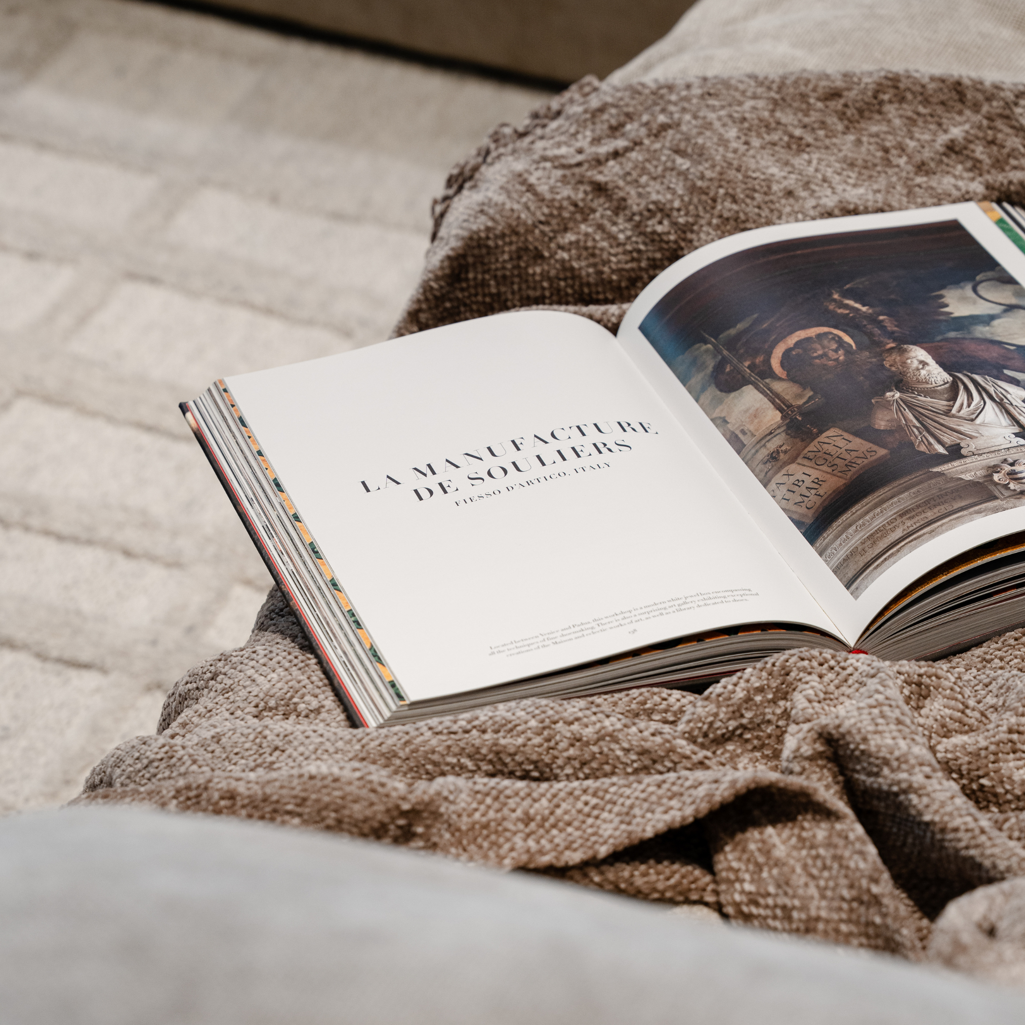 Close-up of an open book resting on a textured beige fabric surface, showing illustrated pages with detailed artwork.