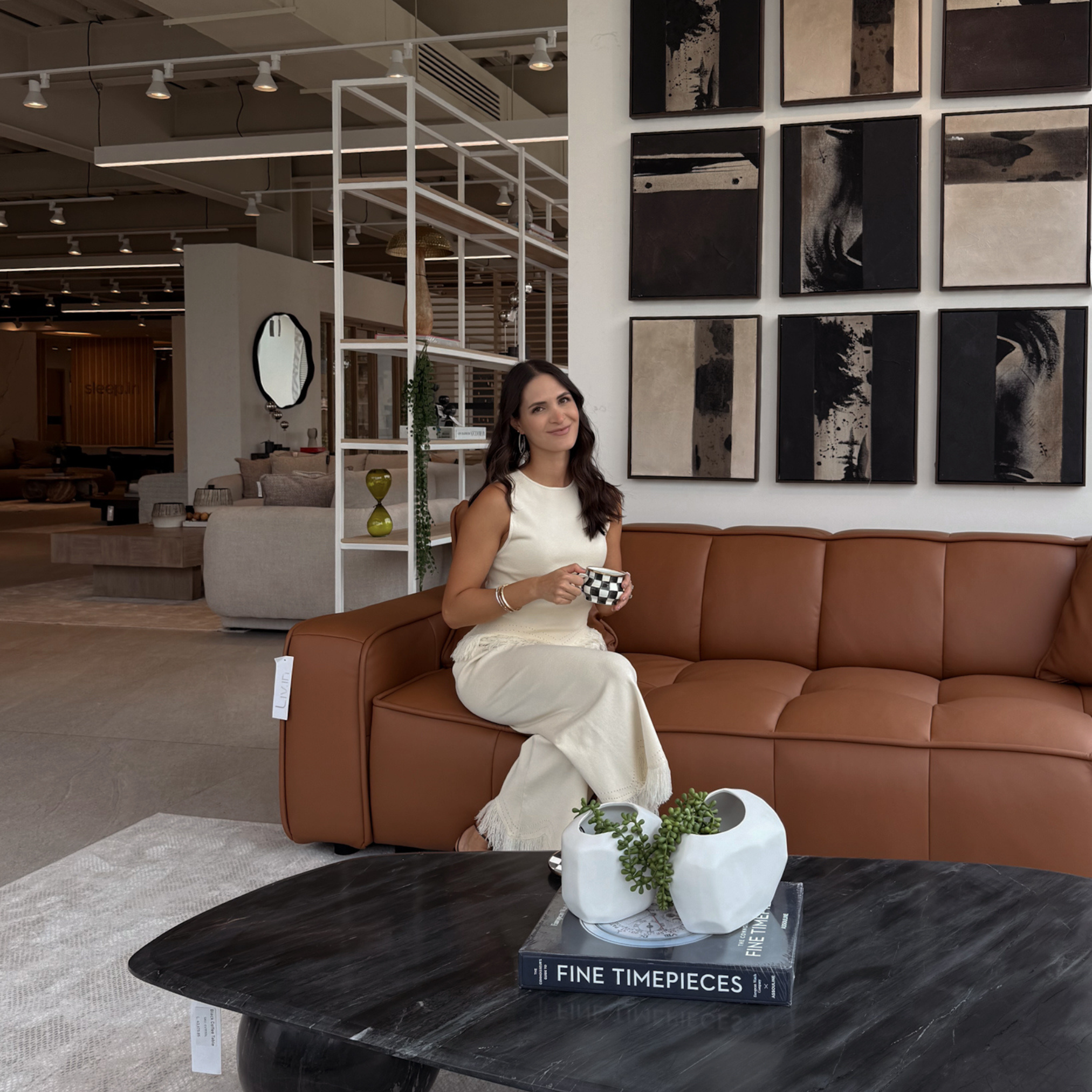A woman in a white dress sitting on a brown leather sofa in a modern interior, holding a black and white checkered cup, with a concrete wall decorated with abstract black, beige, and brown artwork, a white shelving unit with decorative objects, and a