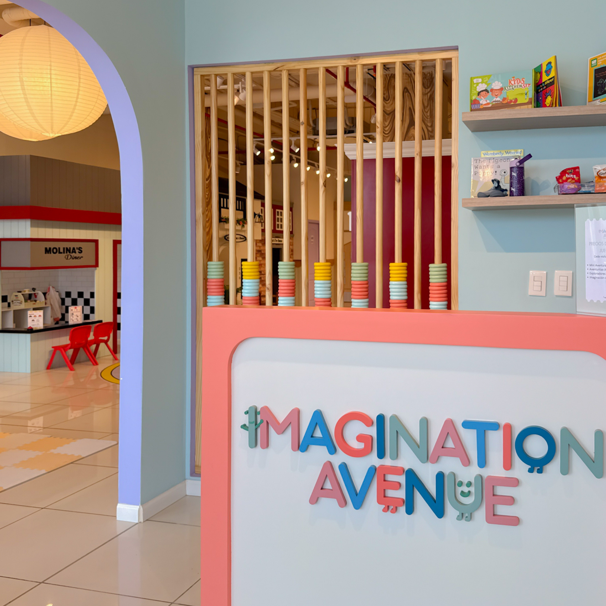 Colorful sign on the wall saying 'IMAGINE, CREATE, ADVENTURE' with a playful rainbow and smiley face design, inside a children's play area or museum.