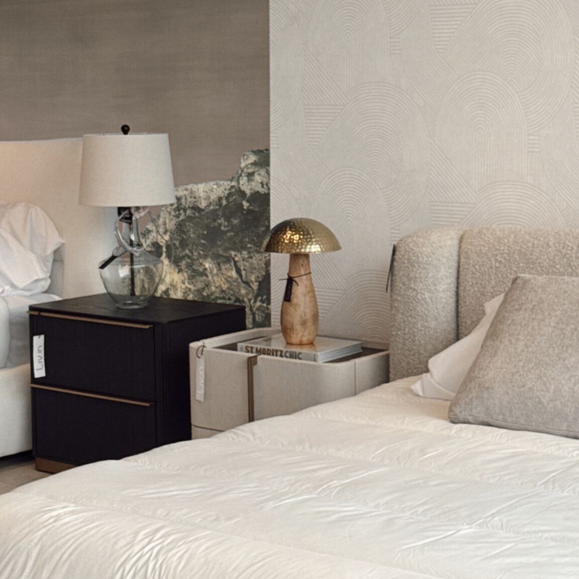 A modern bedroom scene featuring a bed with white linens, a black nightstand with a glass vase, a white table lamp, a stone sculpture, and a gold table lamp on a white side table, with a textured geometric wallpaper background.