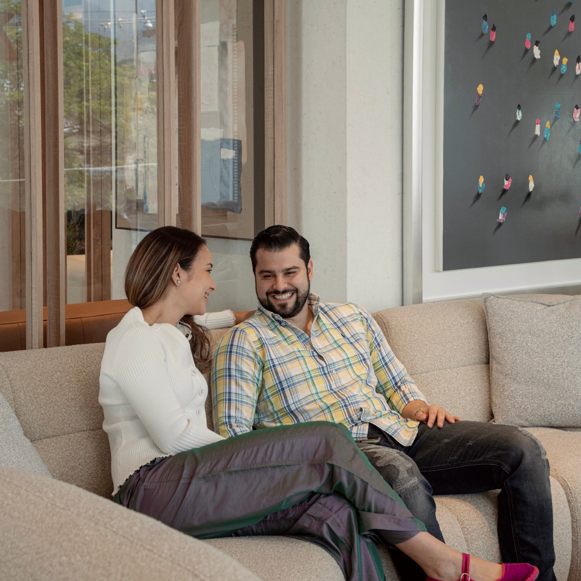 A woman and a man sitting on a beige couch talking and smiling, in a living room with wood accents and a decorative board with colorful pinned items on the wall.