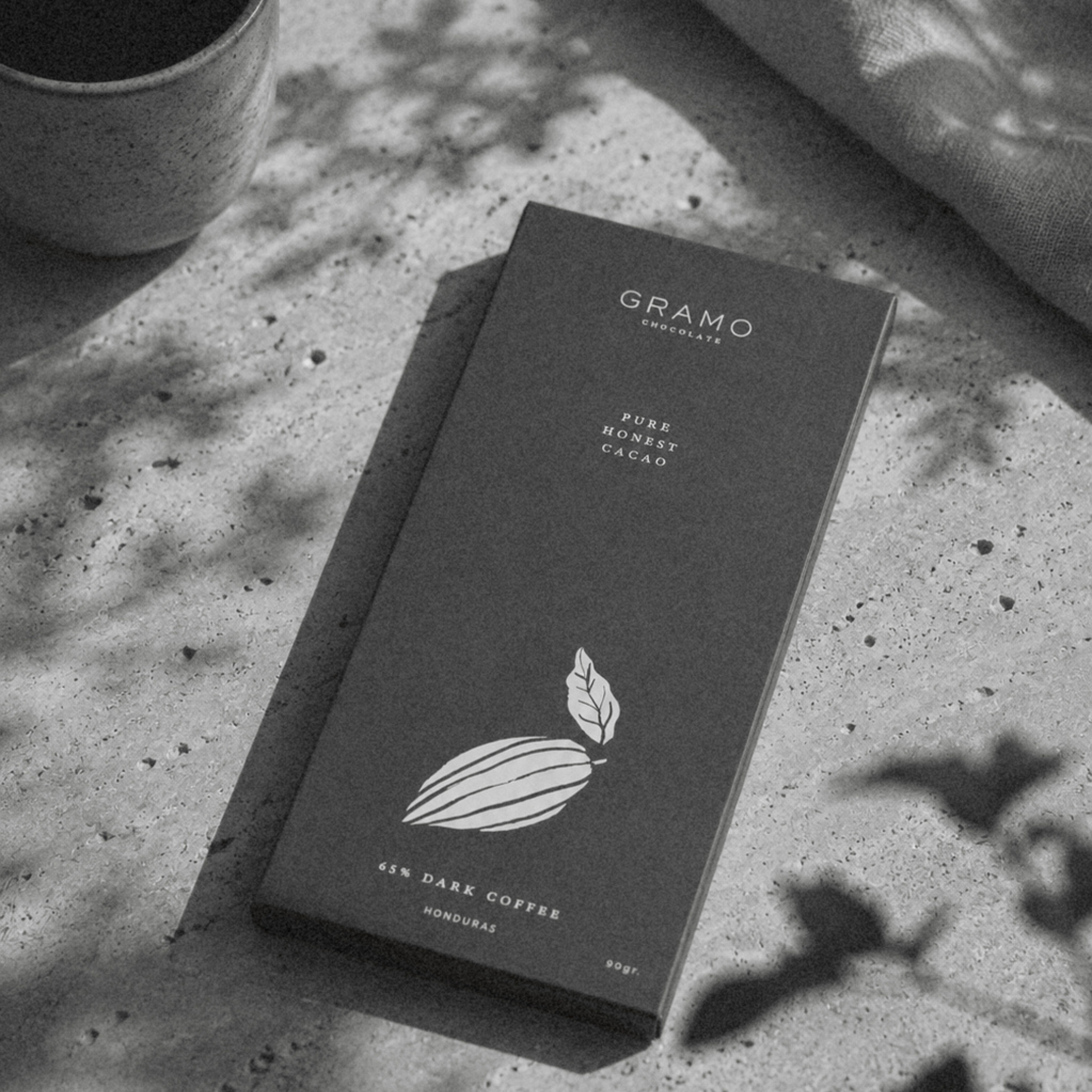 A black box of Gramo chocolate with a leaf logo on a concrete surface, with a mug of coffee and a paper napkin nearby.