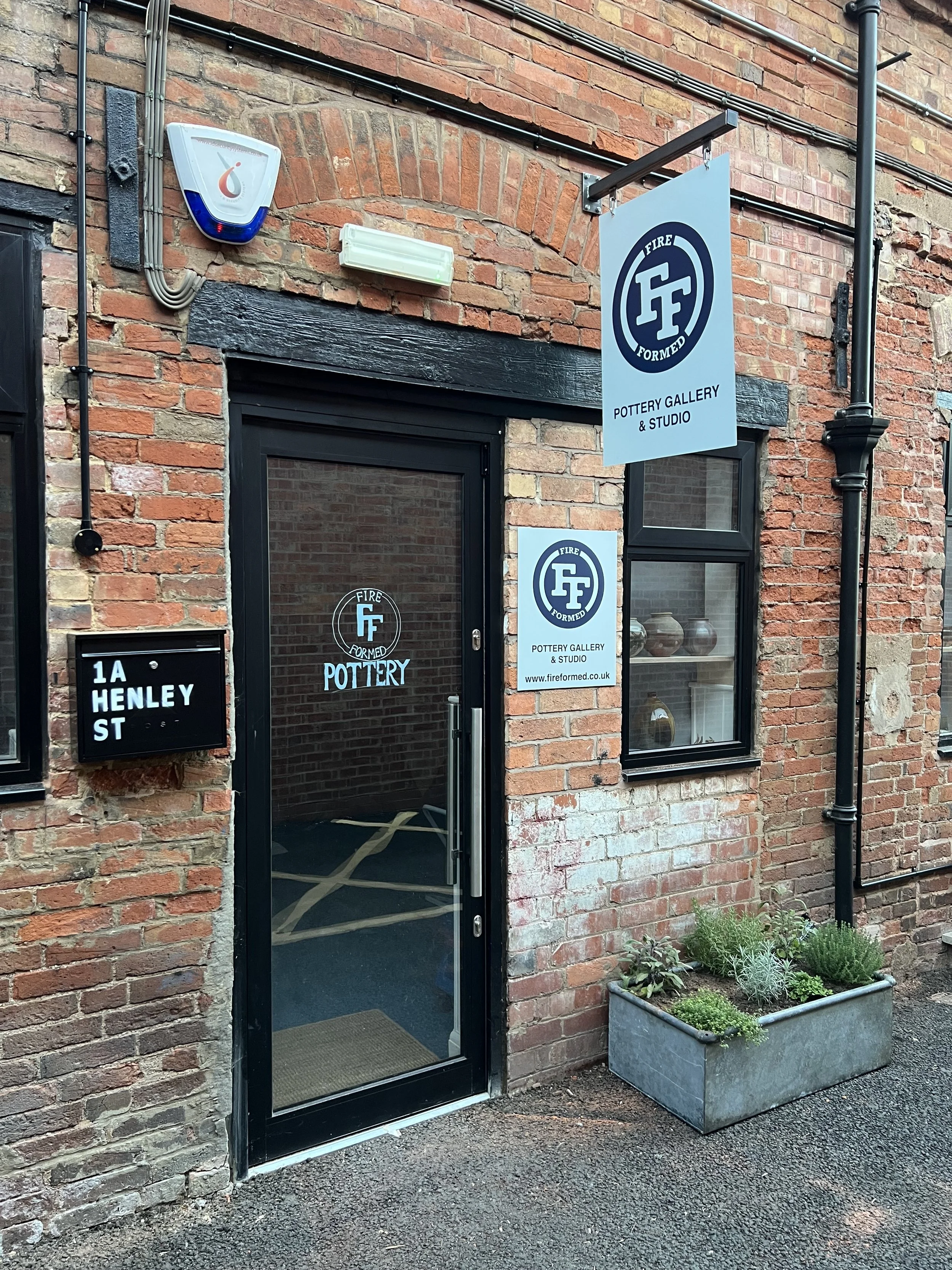 Brick building entrance with black door and window, sign reading 'Pottery Gallery & Studio,' a black street number plaque '1A Henley St,' a small garden with green plants in a rectangular gray planter, and a sidewalk.