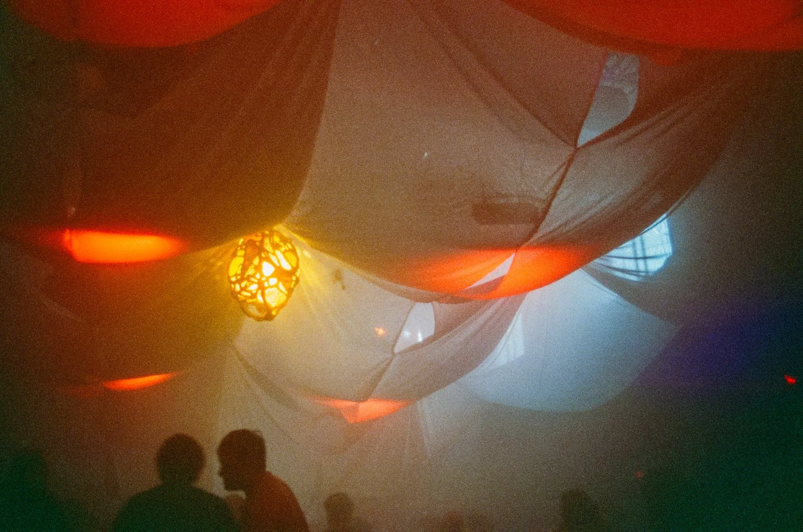 Colorful ceiling installation with balloons and hanging lights, showing silhouettes of people in a dimly lit room.