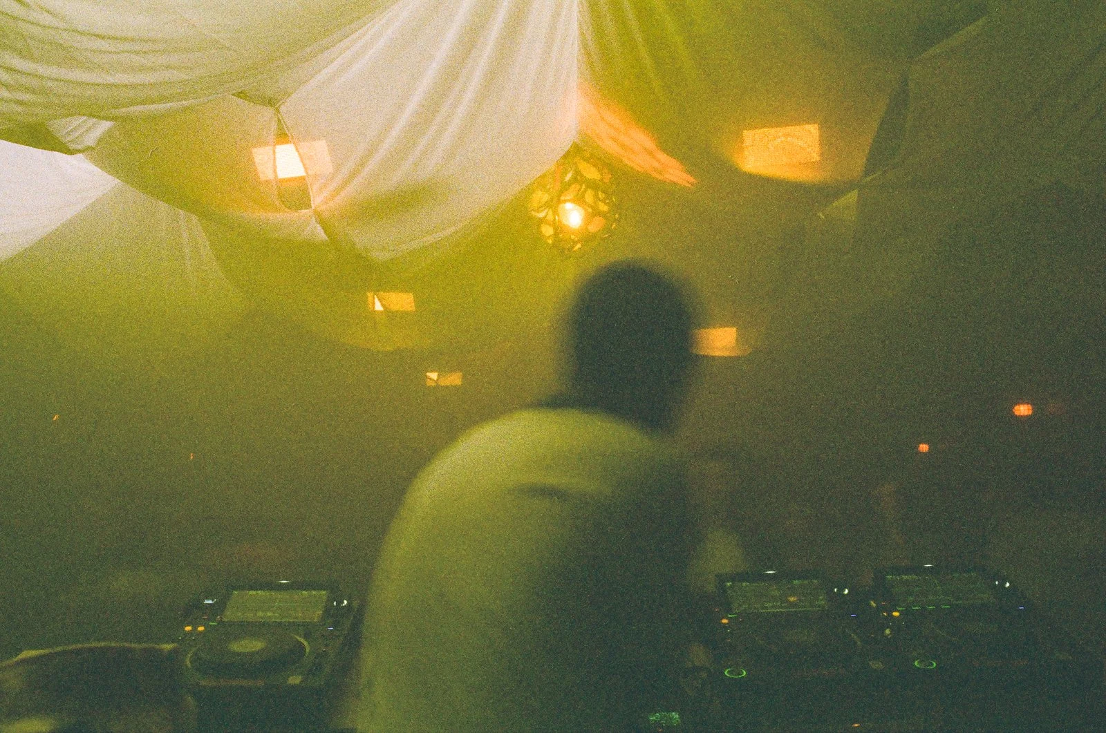 A person with a blurred silhouette standing behind DJ equipment in a dimly lit setting with hanging fabric and warm lighting.