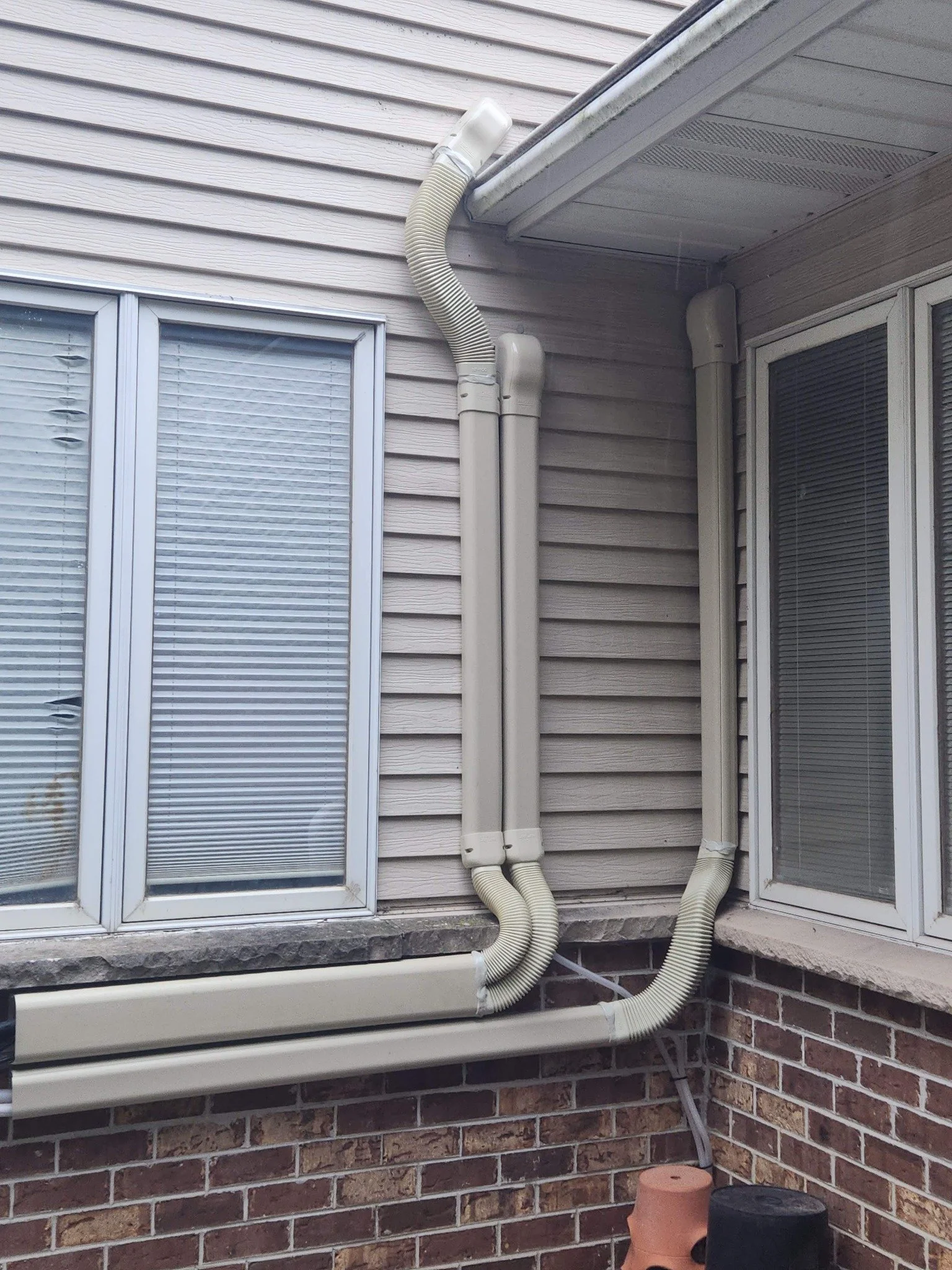 Exterior view of a house showing two windows with blinds, beige siding, and white downspouts with flexible extension hoses attached to the gutter system.