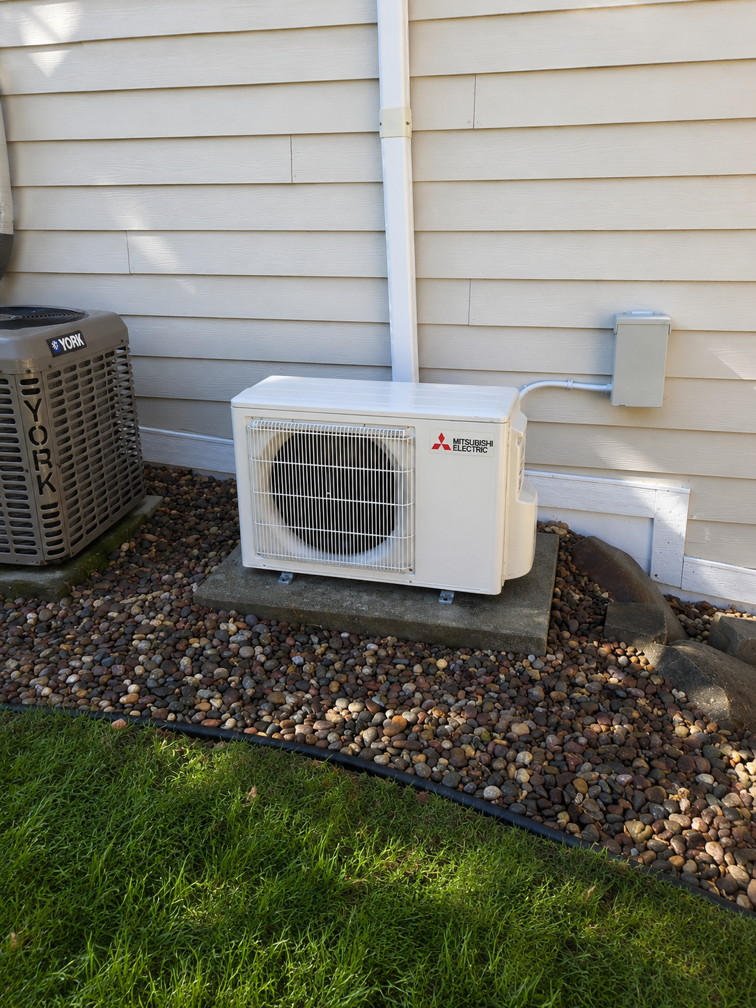 An outdoor air conditioning unit with Mitsubishi Electric branding installed on a concrete slab near a house with vinyl siding, alongside another HVAC unit on a gravel ground with grass nearby.
