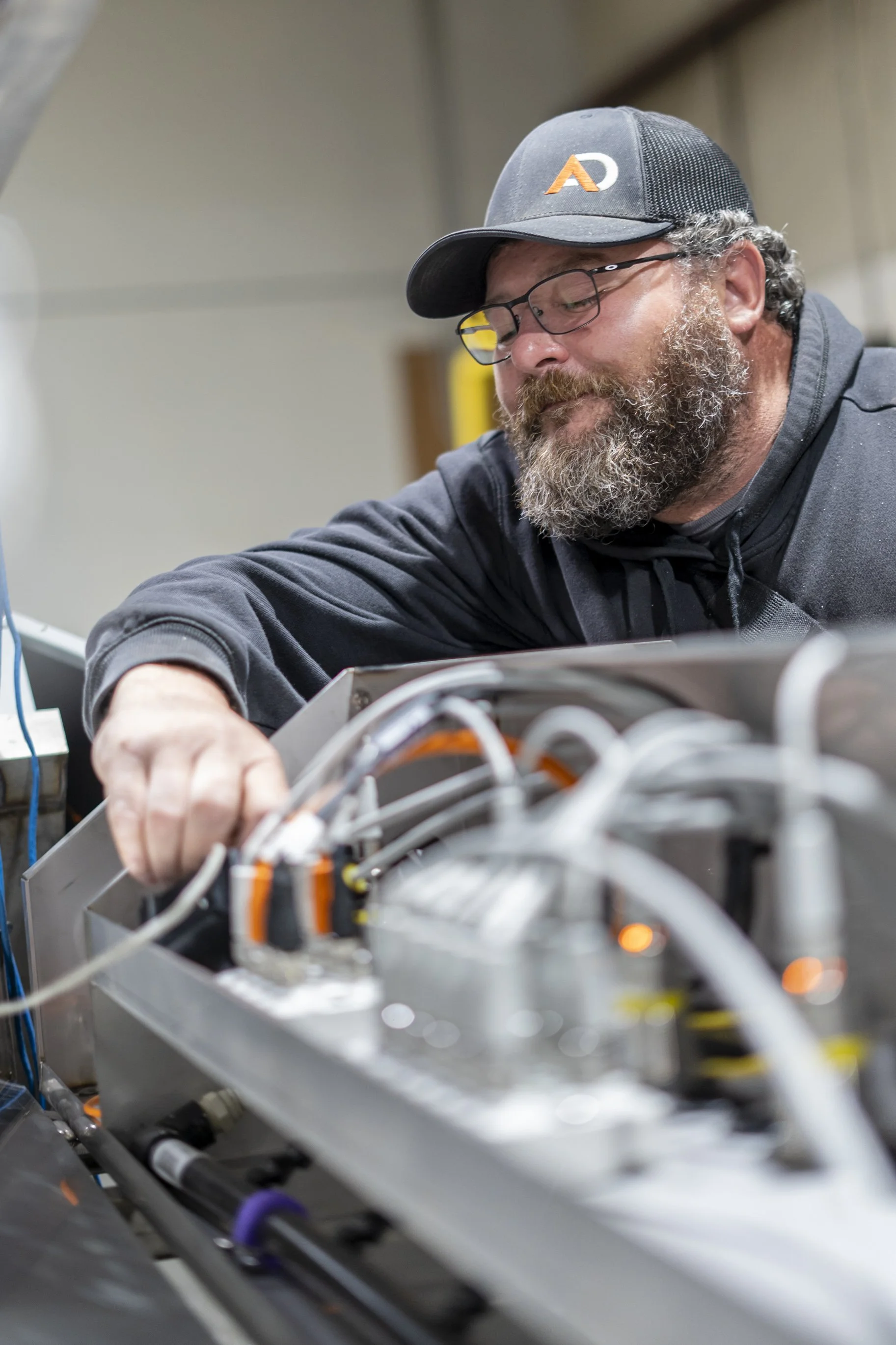 A man with glasses and a beard wearing a black cap is working on dairy electronic equipment with numerous wires and circuit boards.