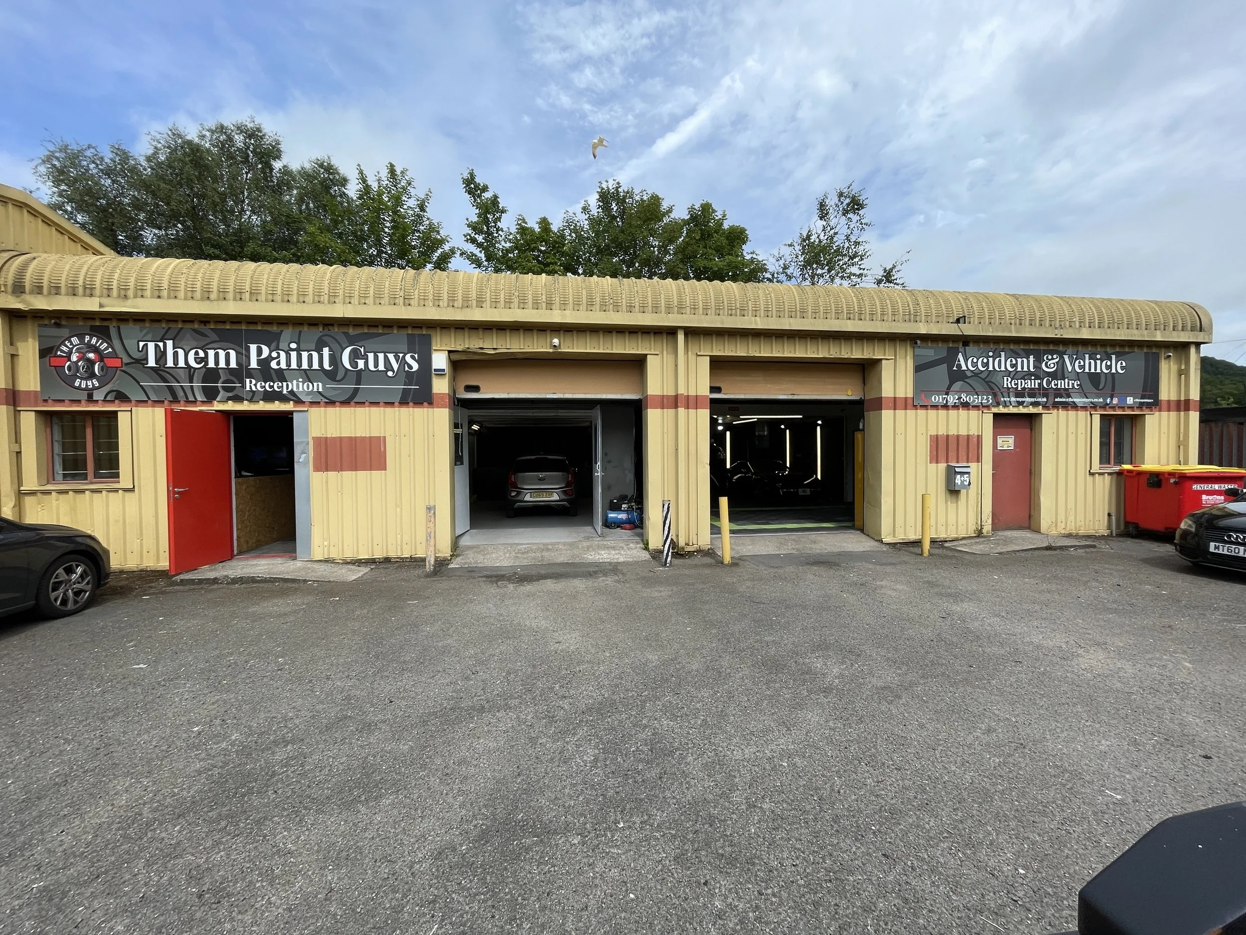 Exterior view of Them Paint Guys, an auto repair shop with a beige and red building including a reception area and garage bays. There is a red trash bin outside and cars parked in front.