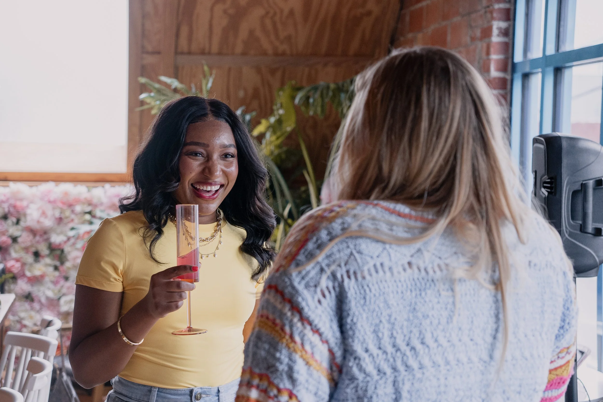 Two women talking at an indoor event, one holding a pink drink, in a room with exposed brick and large windows.