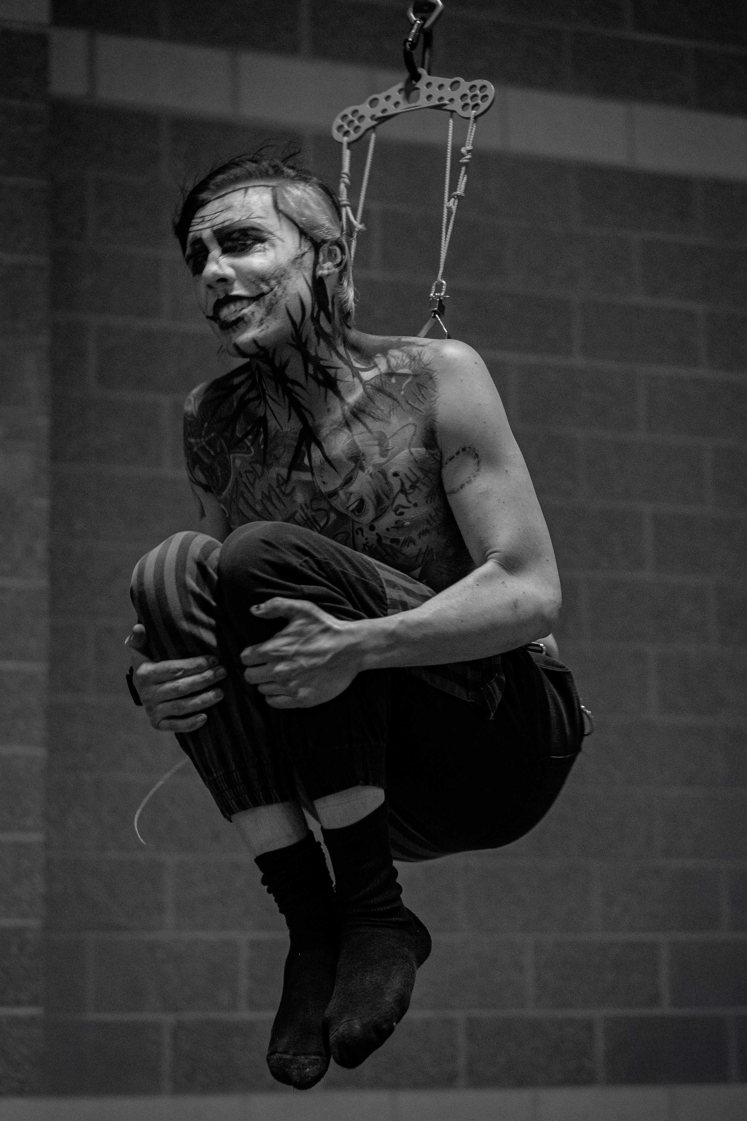 Black and white photo of a person hanging upside down from a harness, with face paint and tattoos, in an indoor setting.