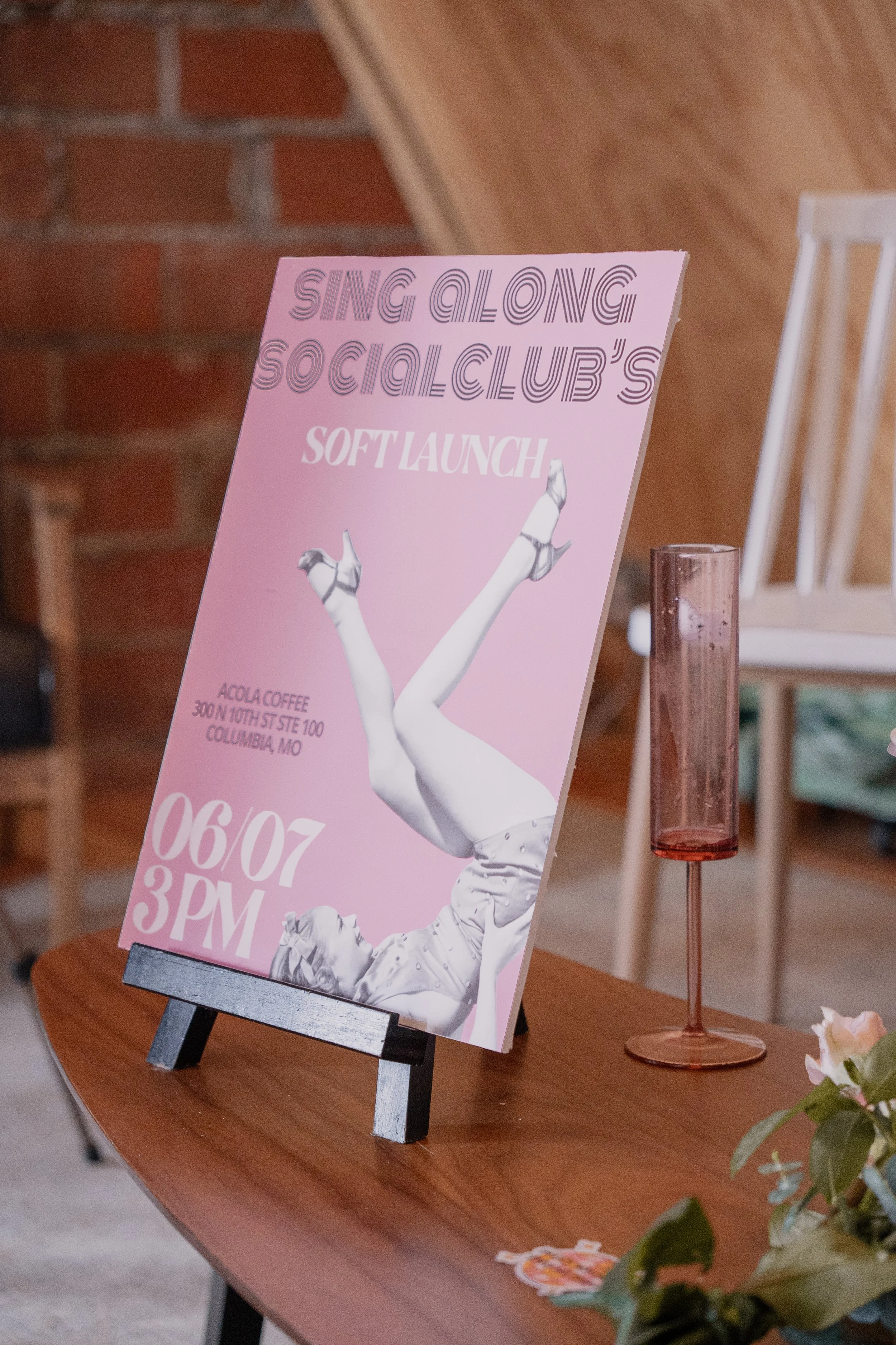 Pink poster on a wooden table with black easel advertising singing and social club's soft launch, with a person singing, and location details at the bottom. Nearby is a pink transparent glass and some flowers.